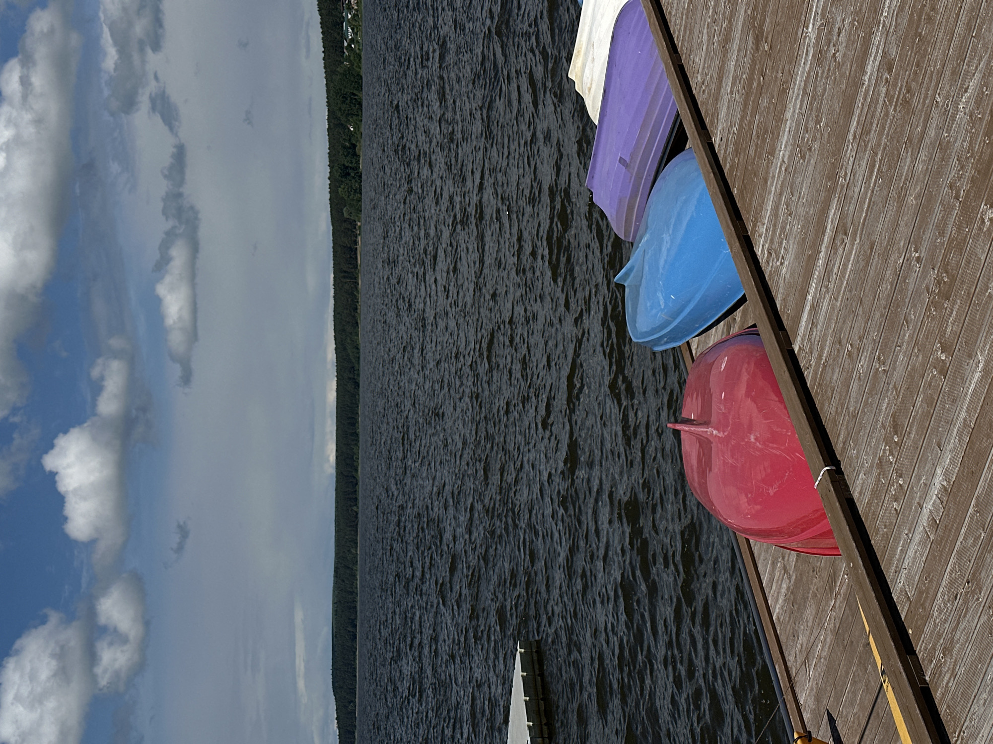 The scene depicts a view from a wooden dock extending into a large body of water. On the dock, three upside-down, colorful boats are resting: one red, one blue, and one lavender/white. Beyond the water, a dense line of trees is visible along the shoreline. In the sky, there are clouds and patches of blue. The location is Votkinsk, Russia.