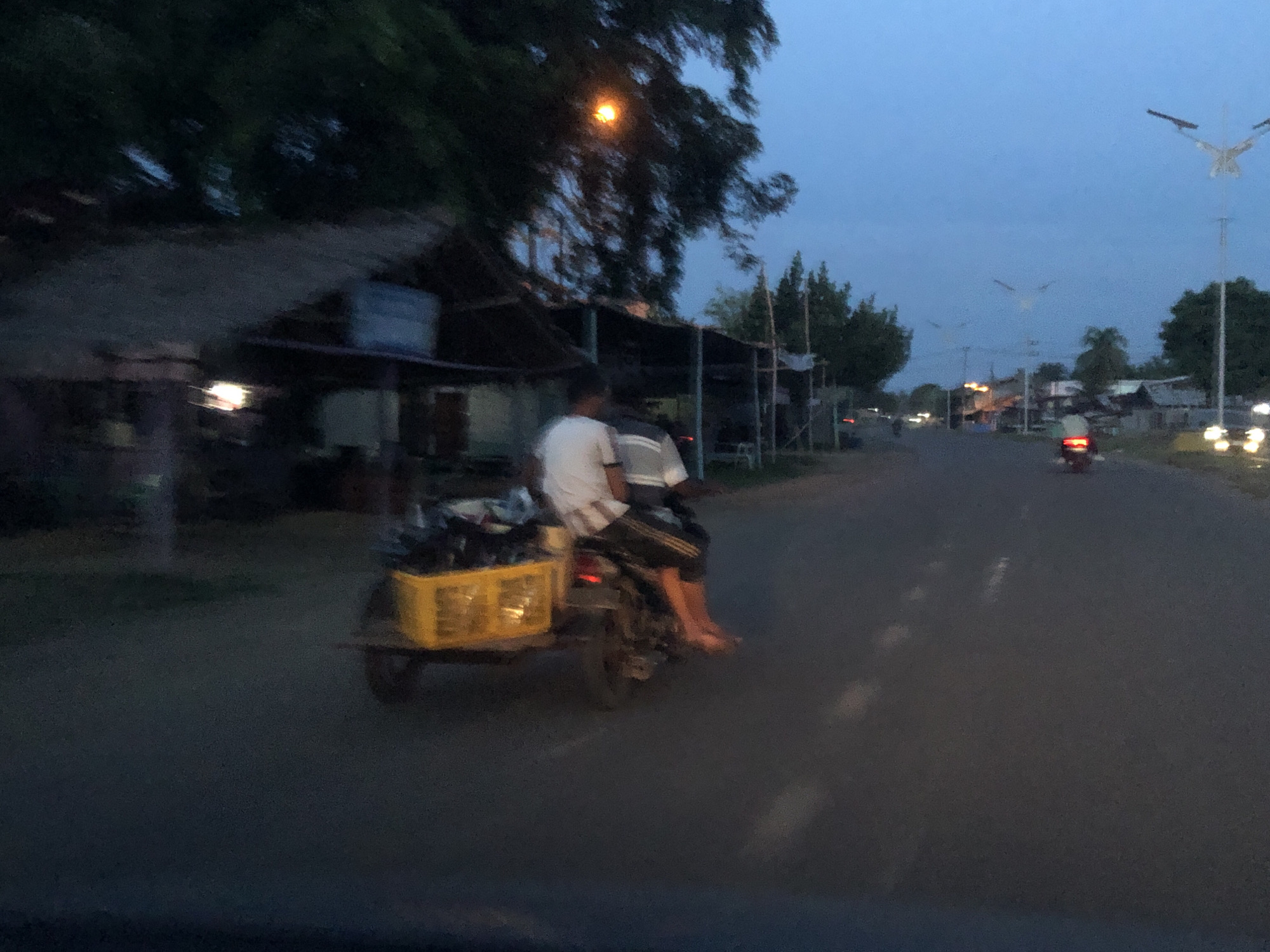 The image captures a street scene at dusk. A motorbike with a trailer attached to the back is moving along the road. Two people are on the motorbike. The trailer contains a yellow container with objects inside. The road is asphalt, and there are markings visible. Buildings with makeshift roofs line the left side of the road. Streetlights are visible in the distance and along the right side of the road. Another motorbike is further down the road. The sky is blue.