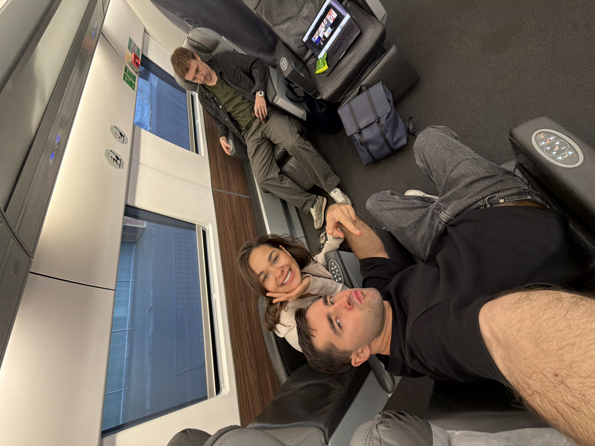Three people are in a train compartment. A man in a black jacket and olive green shirt, and brown pants is reclined in a chair. On the adjacent seat are a laptop computer, a blue bag and a snack food. A woman sits holding the hand of a man who is lying down, seemingly taking a selfie. The train appears to be moving in an urban area, judging by the buildings seen out the window. Location: likely Tbilisi, Georgia, but verification would require additional identifying information.