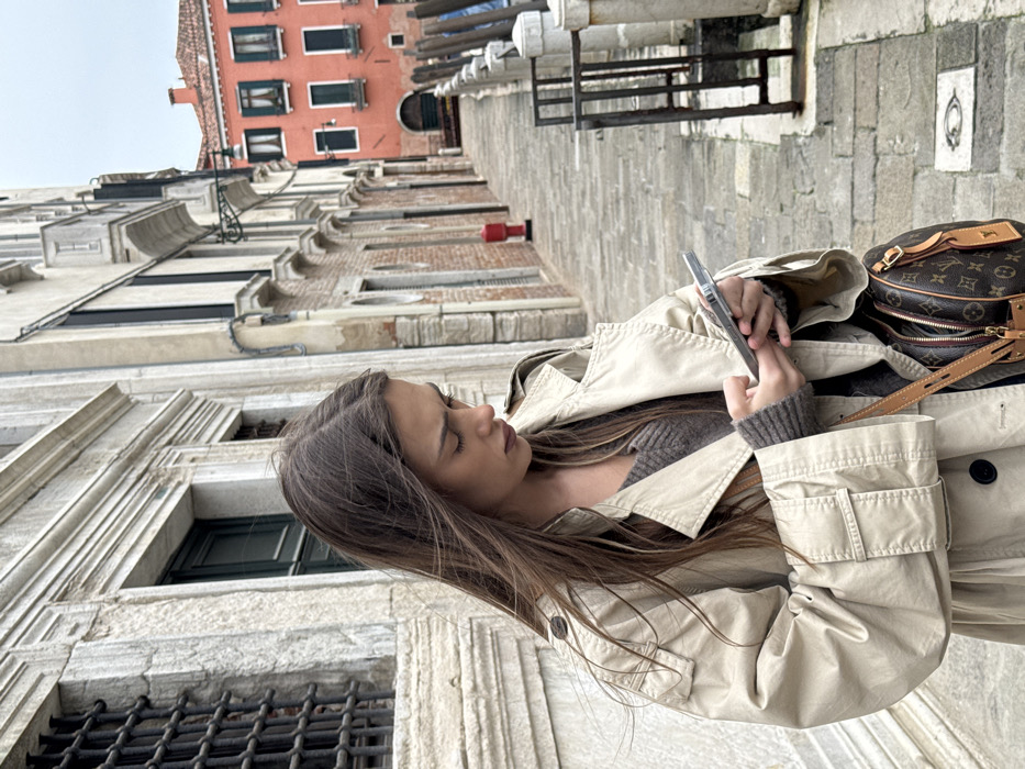 A young adult female stands on a paved stone walkway in Venice, Italy, viewed from the mid-torso up. She has long, dark brown hair and wears a light beige trench coat over a grey V-neck sweater. A brown Louis Vuitton shoulder bag hangs on her right side. Her gaze is directed downwards, focused on a silver-colored smartphone she holds horizontally with both hands, appearing to interact with it.
The immediate environment features a weathered light stone building on the left with a grated window. To the right, grey stone mooring posts line the edge of a canal, indicating a waterside path. The narrow stone walkway recedes into the background, flanked by historic Venetian buildings. A red fire hydrant is visible on a building wall in the mid-distance on the right. Further down the path, a prominent multi-story red-orange building with dark shutters stands out against an overcast sky, characteristic of Venetian architecture.