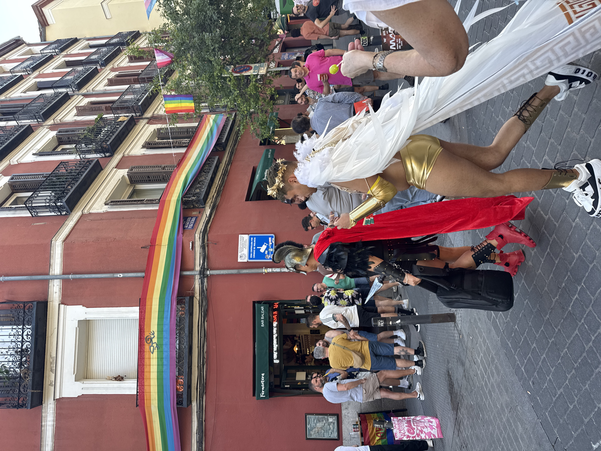 A crowd is gathered on a cobblestone street in Madrid, Spain, outside of a business with a green awning that reads "Fermentera BAR BALEAR". A large rainbow flag is draped across the building's facade. Several people are visible, including some dressed in elaborate costumes. In the foreground, a person dressed as an angel with large white wings and gold attire is walking, while another person in a gladiator-style costume with a red cape is walking beside them. Other individuals in casual clothing are standing and talking. The street is lined with buildings, and there are trees and streetlights visible. A sign on a lamppost indicates "ZONA CONTROLADA POR CÁMARAS DE VIGILANCIA", which translates to "Area controlled by surveillance cameras".
