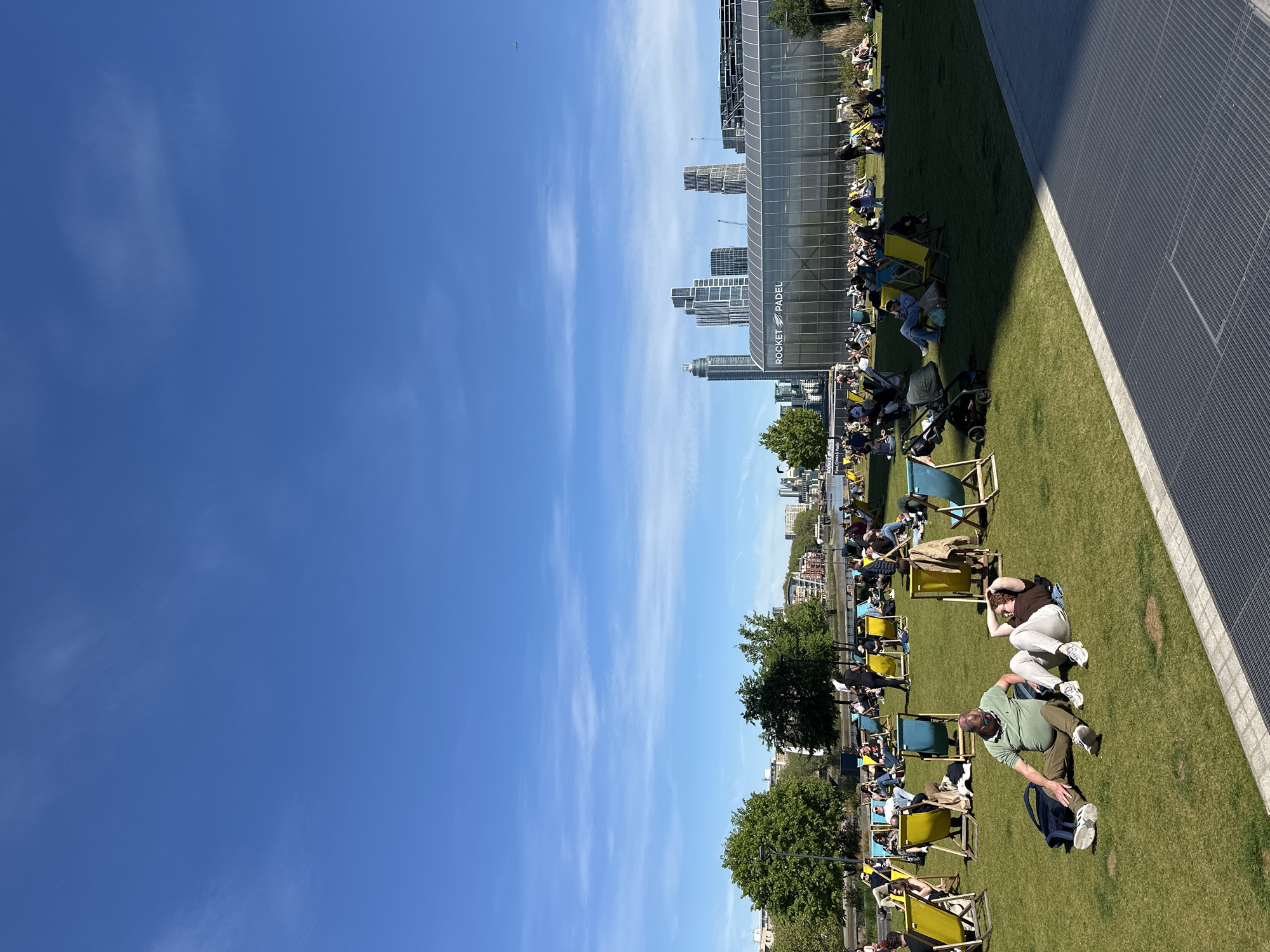 The image shows a sunny day in London, United Kingdom. People are relaxing on the grass, some in deck chairs, at what appears to be a park. In the background, there are modern buildings and skyscrapers. A building with "ROCKET PADEL" written on it is visible. The sky is blue with some clouds. There are trees and greenery interspersed among the people.