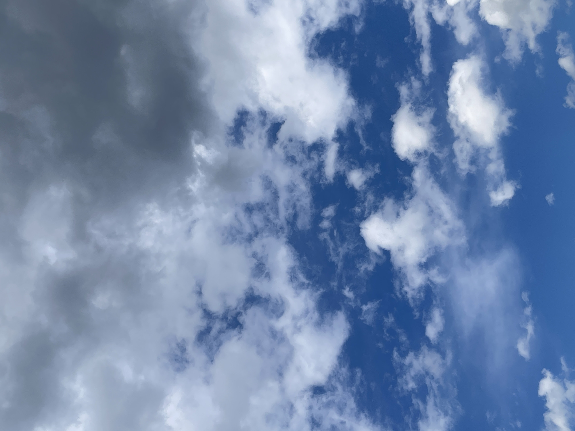 The image shows a cloudy sky, with a mix of dark, heavy clouds on the left side and scattered white clouds against a blue sky on the right. The dark clouds appear to be storm clouds, while the white clouds are more typical cumulus clouds. There is no information about the location in the city of Unknown, Unknown, or any people or subjects.