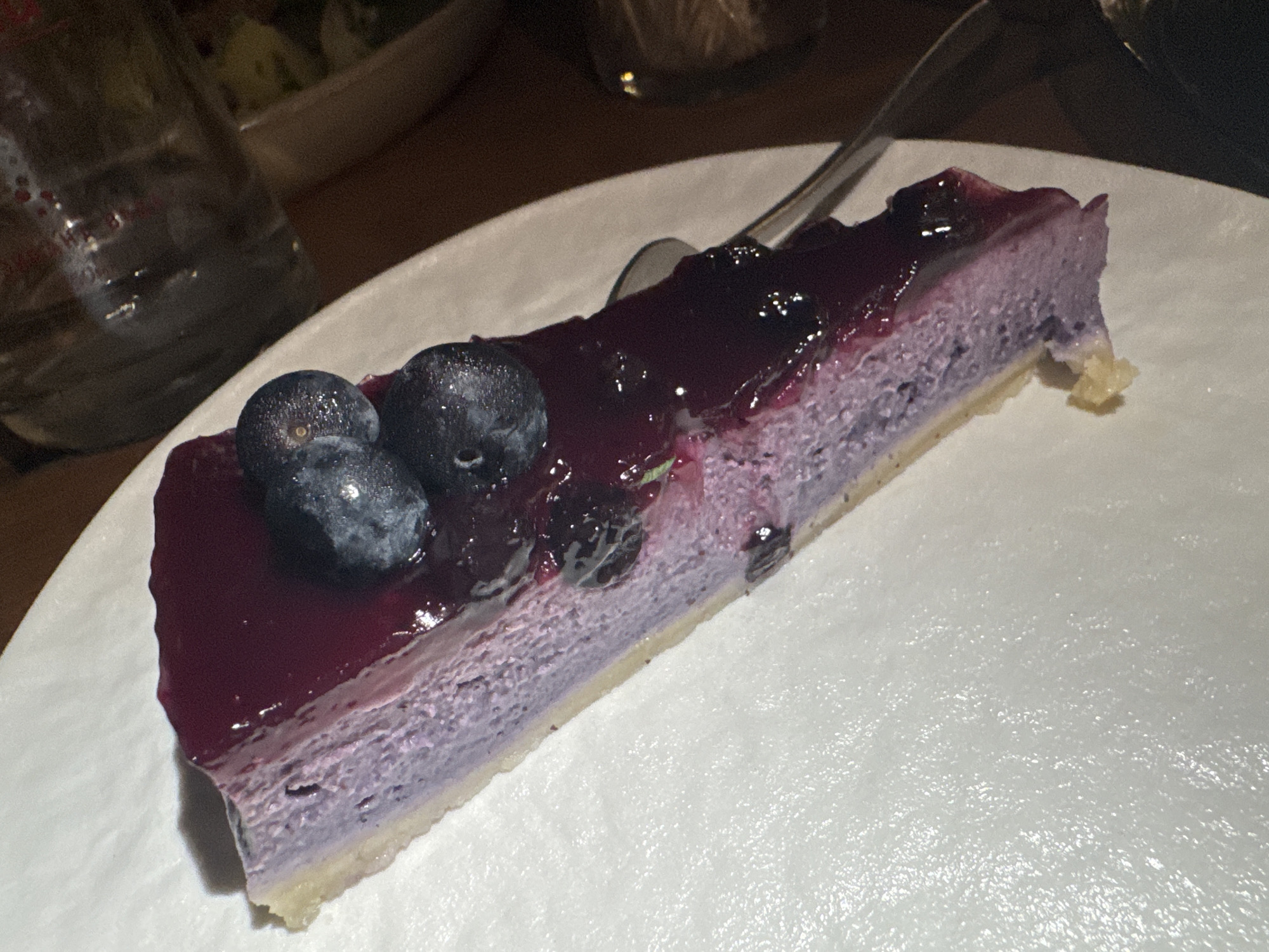A single wedge of blueberry cheesecake is positioned diagonally on a plain white, subtly textured round plate. The dessert features a distinct, pale gold crust base, surmounted by a thick layer of pastel purple cheesecake filling that exhibits a slightly granular texture. A rich, dark purple blueberry compote, glossy and reflecting ambient light, covers the top surface of the cheesecake, containing visible pieces of fruit. Three fresh, plump blueberries, some with tiny water droplets, are arranged on the compote at the wider end of the slice. A silver eating utensil, likely a spoon, is partially visible behind the dessert, resting on the plate. The background is significantly blurred and dark, revealing the upper portion of a clear glass bottle or carafe on the left, and a very indistinct, light-colored object, possibly another dish, further back. The environment suggests an intimate indoor dining setting, such as a restaurant or cafe located in Plovdiv, Bulgaria.