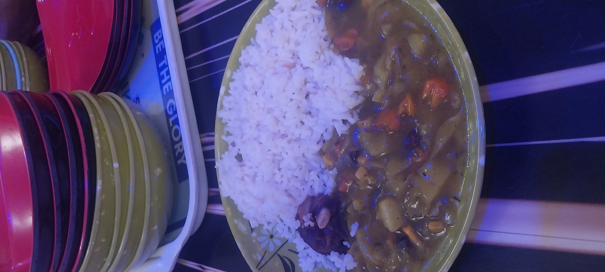 In the Akpaka Forest Reserve, Nigeria, a table is set with a meal. The focus is on a green plate containing white rice and a stew. The stew appears to be a vegetable and meat curry, with visible pieces of carrots, potatoes, and a chunk of dark meat. The plate is positioned on a black and white striped placemat. In the background, a stack of colorful bowls, primarily red and green, are piled high. A white tray is visible beneath the bowls, with blue lettering that reads "BE THE GLORY". The lighting in the scene is dim with noticeable blue and purple hues, possibly from ambient lighting or a flash.
