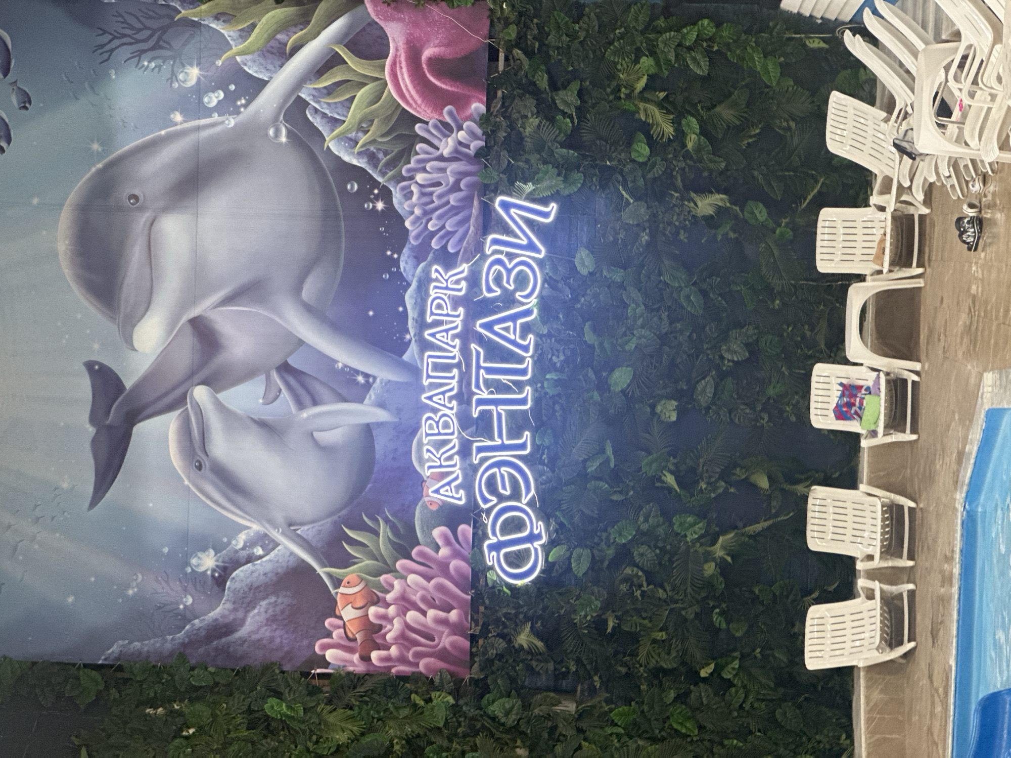 The image shows an indoor water park with a large mural on the wall depicting dolphins and marine life. Neon signs above the mural spell out "АКВАПАРК ФЭНТАЗИ" in Russian, which translates to "FANTASY WATER PARK". The water park appears to be designed with a tropical theme, featuring artificial greenery and a pool in the foreground. Several white lounge chairs are arranged near the pool. This water park is located in Moscow, Russia.