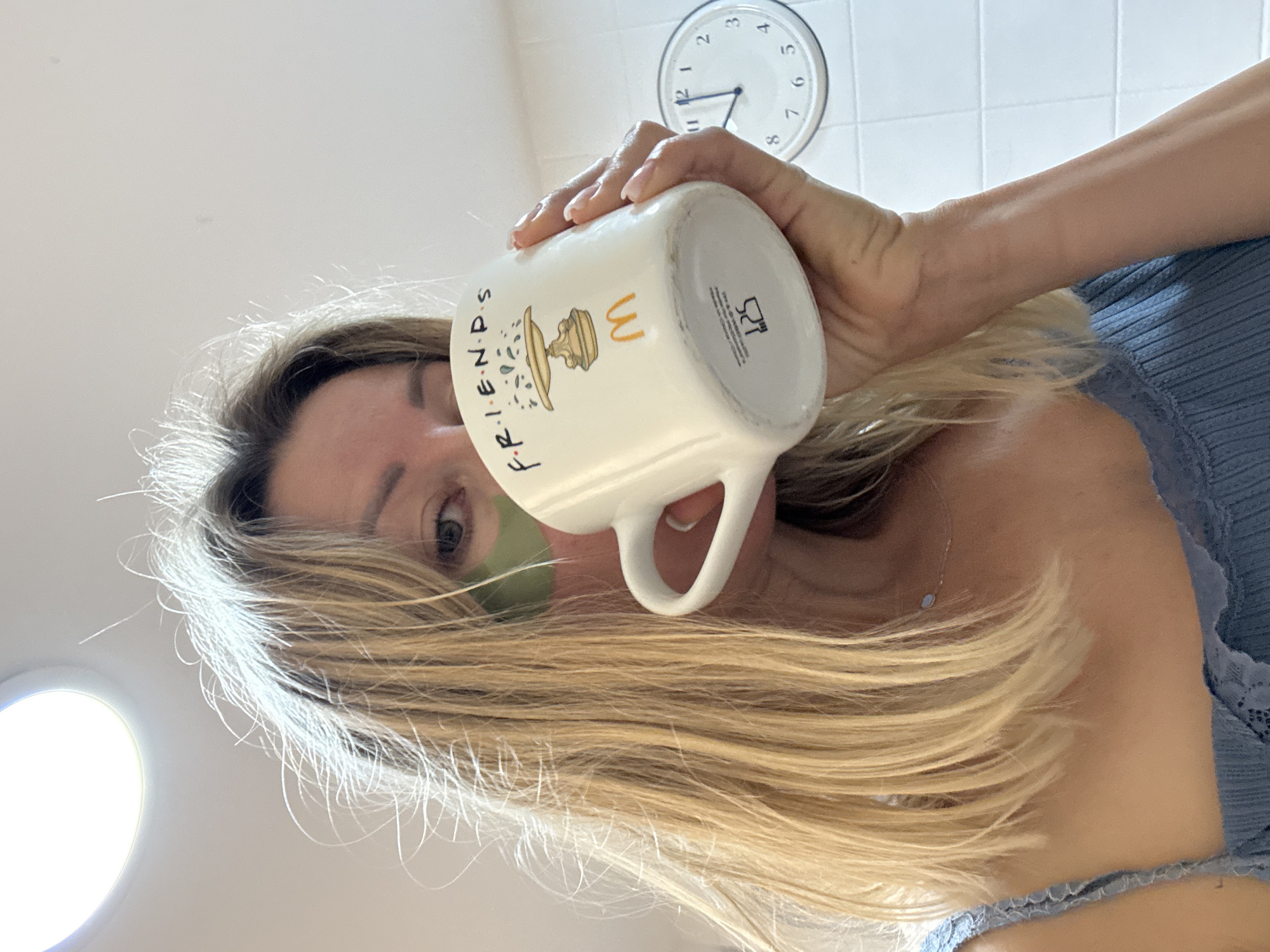 A woman with blonde hair and green eye patches is holding a white mug with the "Friends" logo and a McDonald's logo. She is drinking from the mug and looking at the camera. The background is a tiled wall with a clock showing the time as approximately 2:52. A bright light is visible in the upper left corner of the image. The image appears to have been taken indoors, likely in a kitchen or bathroom.