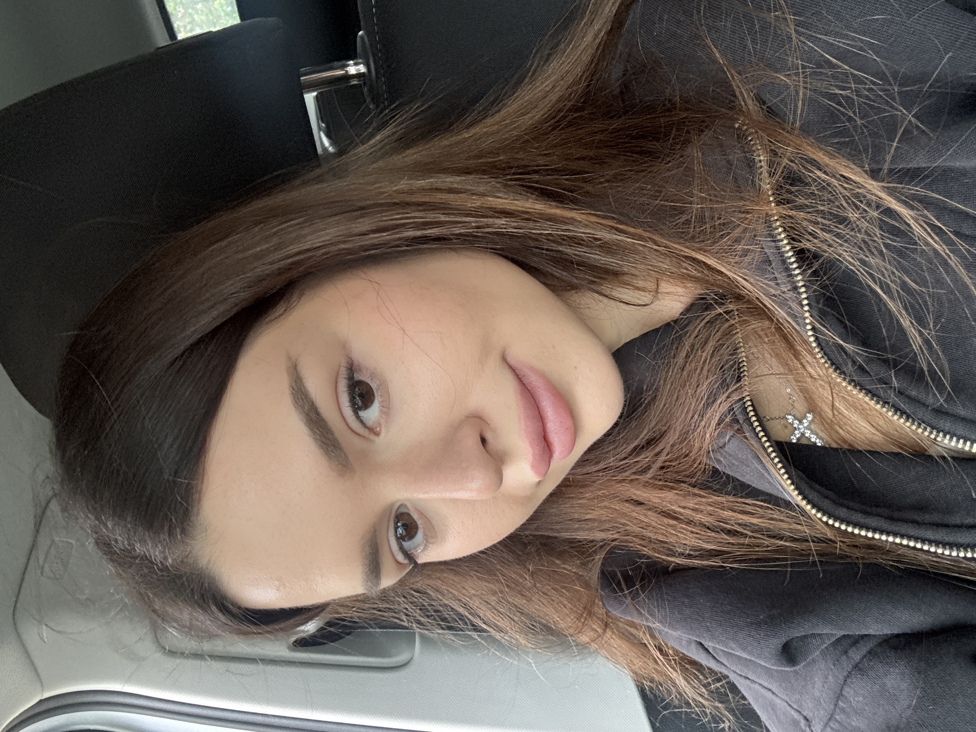 The image shows a young woman with long, brown hair, sitting in the driver's seat of a car. She is wearing a dark-colored zip-up jacket and a necklace with a cross pendant. Her eyes are brown, and she has full lips. The interior of the car is visible, including the headrest and part of the dashboard. The lighting suggests it is daytime. The image does not provide any information about the location being Prague, Czechia.