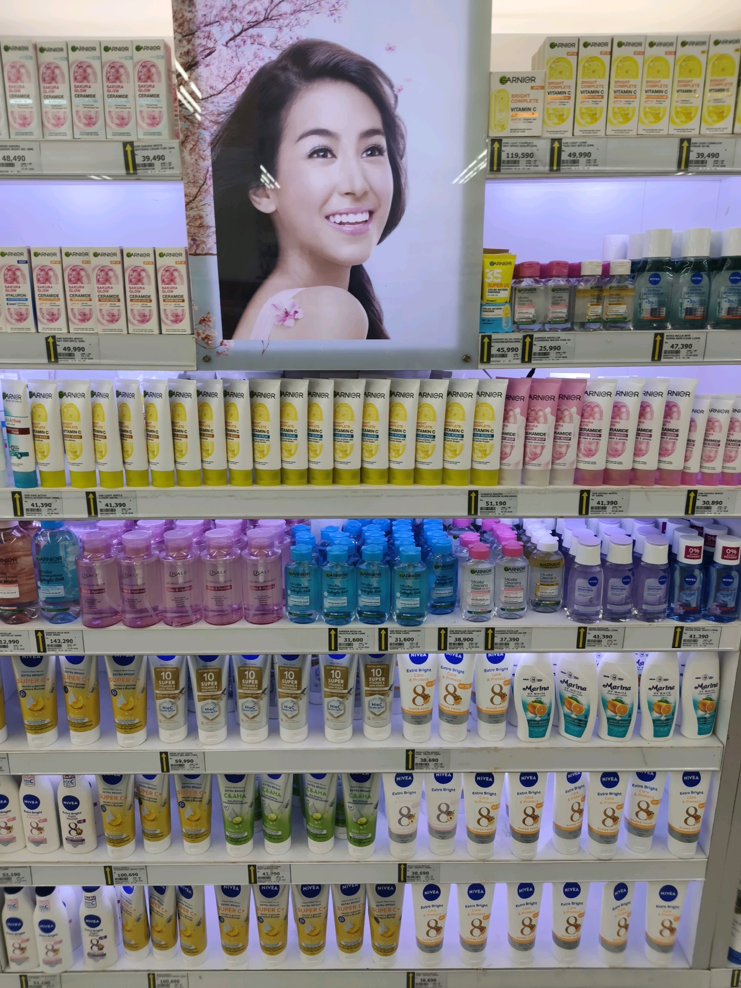 The image depicts a retail display of skincare products in a brightly lit store, likely a supermarket or drugstore. The display features multiple shelves stocked with various lotions, creams, micellar waters, and facial cleansers from brands such as Garnier and Nivea. Many products are arranged in rows and packaged in colorful tubes and bottles. In the background, a large promotional poster of a smiling woman, likely a celebrity endorser, is visible, with cherry blossom imagery suggesting a theme of beauty and freshness. Pricing tags are present on some shelves, indicating specific product costs. The environment appears clean and organized, typical of a retail setting. The presence of Indonesian Rupiah pricing (indicated by the comma usage in numbers like 48,490 and 39,490) strongly suggests the location is within Indonesia. Given the context of retail displays and product availability common in Indonesian supermarkets, a plausible location for such a scene in the city of Ketapang, Indonesia, would be a modern supermarket or a large department store.