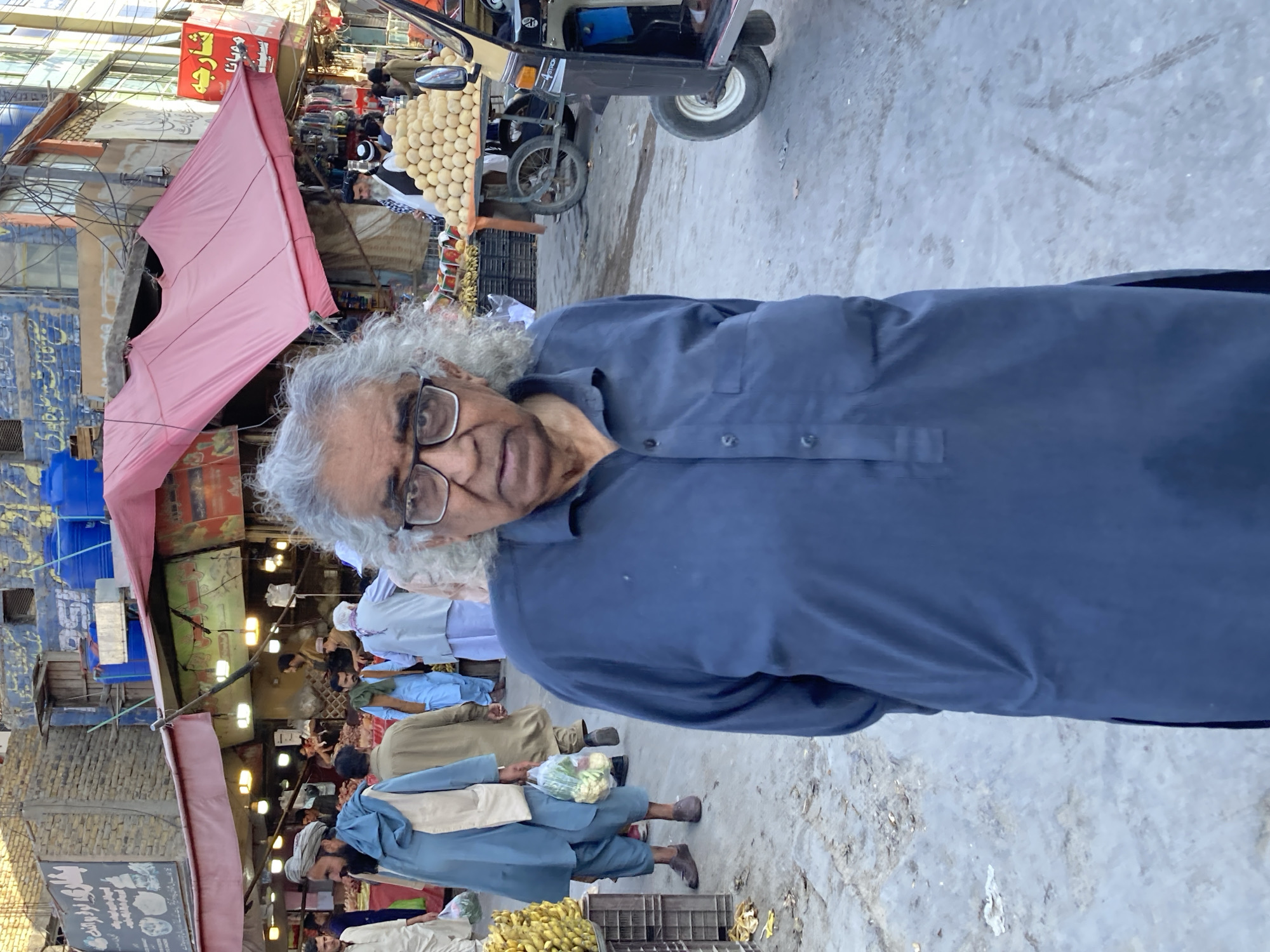 This image captures a bustling outdoor market scene in Quetta City Tehsil, Pakistan, during daylight hours. The central subject is an older man with distinctive grey, curly hair and eyeglasses, wearing a dark blue kurta. He is looking directly at the camera with a neutral expression.
The setting is a busy street or alleyway, with a paved ground that shows signs of wear and dirt. In the background, numerous other individuals, predominantly men dressed in traditional Pakistani attire such as shalwar kameez and kurtas, are seen walking, standing, and engaging in market activities. One man in the mid-ground is holding a clear plastic bag containing vegetables, possibly cauliflower or broccoli.
Market stalls line the street, some identifiable by their prominent pink awnings providing shade. A vendor's cart is visible, laden with a large display of light-colored, spherical items, which could be eggs, sweets, or fruit. Another stall exhibits bananas in crates on the ground. A three-wheeled auto-rickshaw is partially visible in the background, parked near a vendor. Buildings with varied facades, including corrugated metal and painted surfaces, line the street, and blue plastic water barrels are noticeable on one rooftop.
The lighting indicates clear, dry weather. Text in an Arabic script (likely Urdu or Pashto) is visible on several signs in the background, including a red sign and a black one below a pink awning, though the specific words are not clearly legible due to the distance and resolution. The overall atmosphere is one of a vibrant and active daily market.