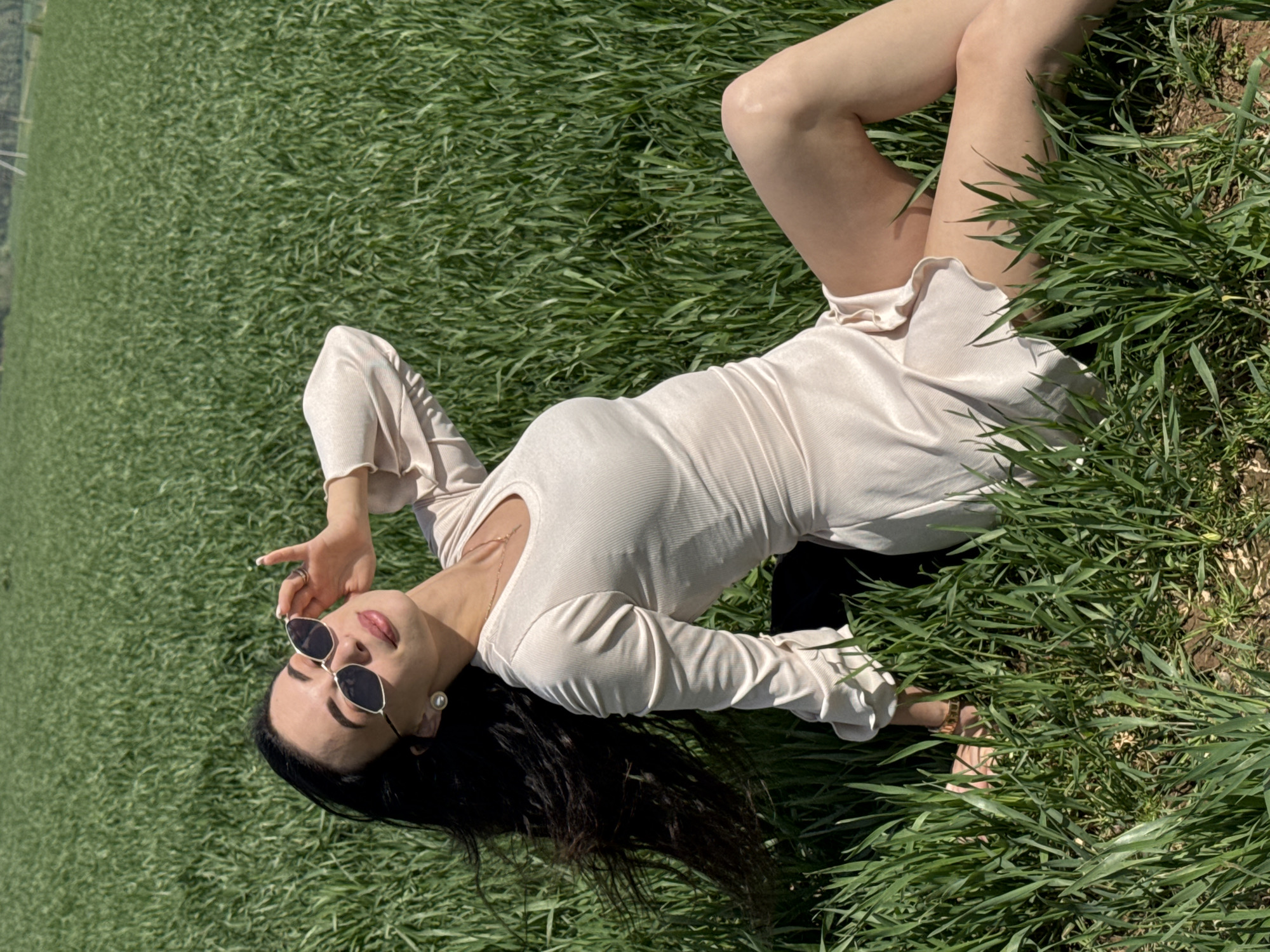 A woman is lying in a field of green grass, wearing a light-colored dress and sunglasses. She is positioned at an angle, with her head towards the bottom of the frame and her legs towards the top. Her left arm is raised, with her hand near her sunglasses. Her dark hair is spread out on the grass. The location is unknown and there is no evidence to indicate this picture was taken in Фоки, Russia.