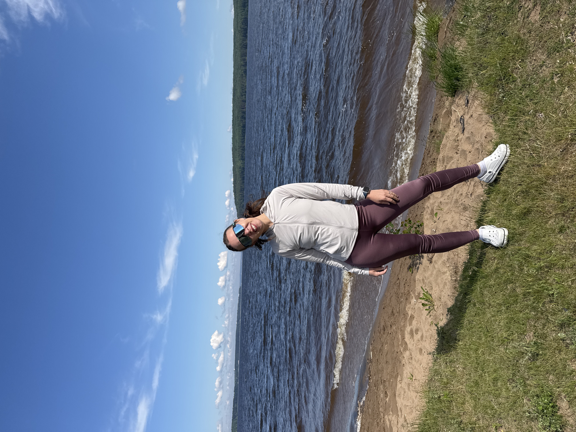 This media file captures a full-body outdoor shot of a woman standing by a large body of water on a bright, sunny day in Chaikovsky, Russia. The image is taken from a slightly low angle, showing the woman positioned between the sandy shore and a patch of green grass.
The woman, with dark hair, is smiling and looking towards the camera. She is wearing a light-colored (possibly light grey or off-white) long-sleeved top with a zipper, dark maroon or purple leggings, white socks, and white Crocs. She also wears reflective blue-tinted sunglasses and a dark-colored watch on her left wrist.
Behind her, a vast expanse of dark blue water stretches out, showing ripples and small waves near the shore. Across the water in the distance, a long, dark green forested landmass is visible, suggesting a large lake or wide river, consistent with the location context of Chaikovsky. The sky above is clear and vibrant blue with scattered wispy white clouds.
The setting appears to be a natural recreational area, with a sandy beach transitioning to lush green grass on the right side of the frame. The bright sunlight, clear skies, and the woman's cheerful expression convey a pleasant and relaxed atmosphere. There is no visible text in the image.