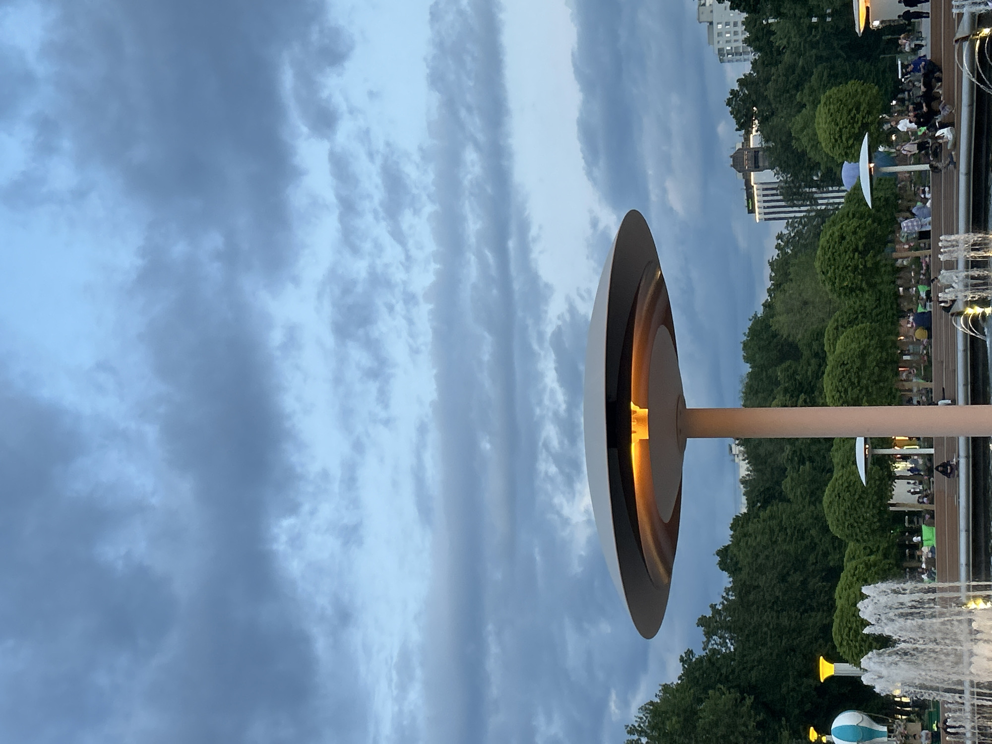 The image depicts an evening scene in a modern urban park or plaza, characterized by artificial lighting against a twilight sky. The sky dominates the upper portion of the frame, filled with textured grey and blue clouds, suggesting dusk or an overcast evening.
In the foreground, a striking contemporary light fixture stands prominently. It features a broad, circular disc-shaped top, beneath which a warm, amber light glows, illuminating its interior. This fixture is supported by a slender, light-colored pole.
To the right, the park area unfolds, populated by numerous individuals and groups. People are seen sitting and standing casually, suggesting a relaxed social atmosphere. Lush green trees and manicured bushes form a dense backdrop, behind which several tall, modern buildings are visible in the distance, indicating an urban setting.
Further to the right, multiple fountains spray water into the air, adding dynamic movement and a refreshing element to the environment. Scattered throughout the park are other smaller, illuminated light fixtures, some with unique designs, including one that resembles a blue and white hot air balloon, enhancing the park's evening ambiance.
The combination of the setting sun's light on the clouds and the activation of artificial lights confirms the time of day as dusk or early evening. The scene conveys a sense of peaceful urban recreation. There is no visible text within the image.