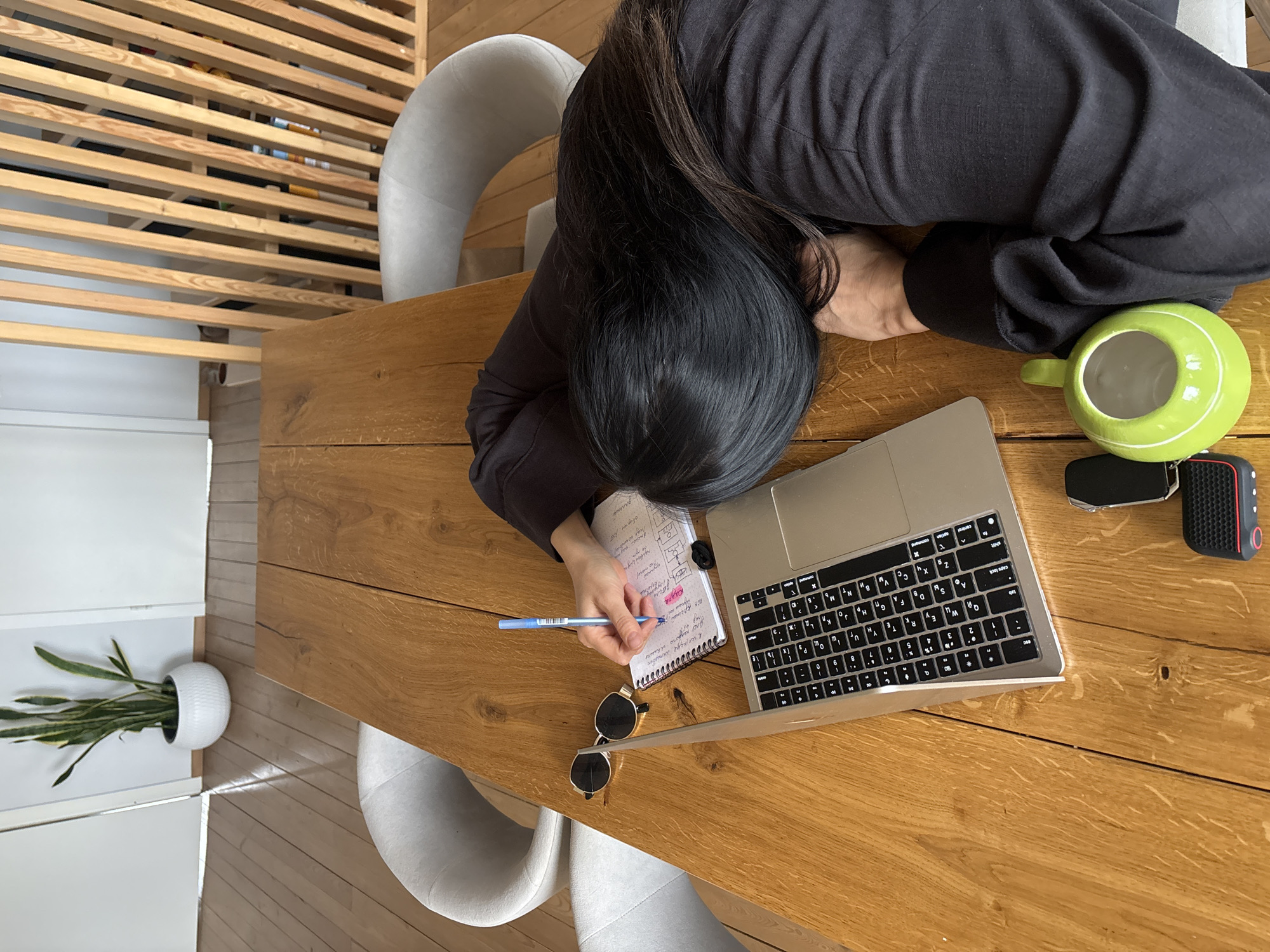 An overhead shot captures a person with long dark hair, wearing a black top, seated at a light wooden table in an indoor setting. Their head is resting on their arm or directly on the table, while one hand holds a blue pen and writes in a spiral notebook.
Arranged on the table are several items: a silver laptop (appears to be a MacBook) with a visible Cyrillic keyboard, a pair of black sunglasses, a bright green mug with a handle, a car key fob, and a small black and red electronic device, possibly a portable speaker.
The background features light wooden flooring, parts of two light grey, curved chairs, and a slatted wooden divider. In the lower left, a white potted plant adds a touch of greenery.
The activity suggests working, studying, or note-taking, with the person's posture possibly indicating deep concentration or fatigue. Notable visible text includes Cyrillic characters on both the laptop keyboard and handwritten notes in the notebook, with the word "Схема" (Scheme/Diagram) clearly legible in the notebook. This detail, combined with the context of Chaikovsky, Russia, strongly suggests the person is in a Russian-speaking environment. The scene is illuminated by natural light, indicating daytime.