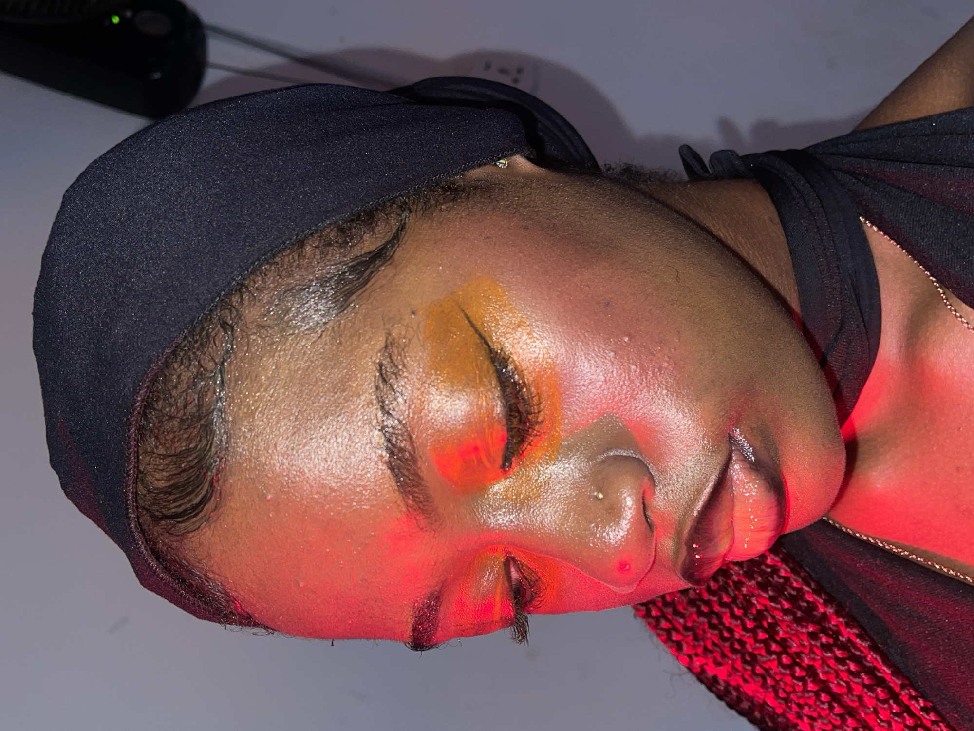 The media file presents a close-up, side-profile portrait of a person with dark skin, located in Ado, Nigeria. The subject's eyes are closed, and their head is slightly tilted back, suggesting a relaxed or posed expression. They are wearing a black headwrap or bonnet, pulled back from the forehead, and a black garment, with a thin gold chain visible around the neck. Red braided hair can be seen peeking from beneath the garment at the bottom right.
The person's face features prominent makeup, including vibrant orange and golden eyeshadow with glitter around the eyes, and dark eyeliner. Their lips appear glossy and full. A small stud piercing is visible on the right nostril. The skin has a dewy or moisturized finish, accentuated by the strong lighting.
The scene is dramatically lit by an intense red light source from the right, casting a warm, crimson glow across the subject's face and upper body. This lighting creates high contrast and highlights facial contours. The background is dim, showing a light-colored wall and a dark, unidentified electronic device with two small green indicator lights in the upper left corner. The setting appears to be indoors, likely at night or in a controlled lighting environment, contributing to an intimate and artistic ambiance. No visible text is present in the image.