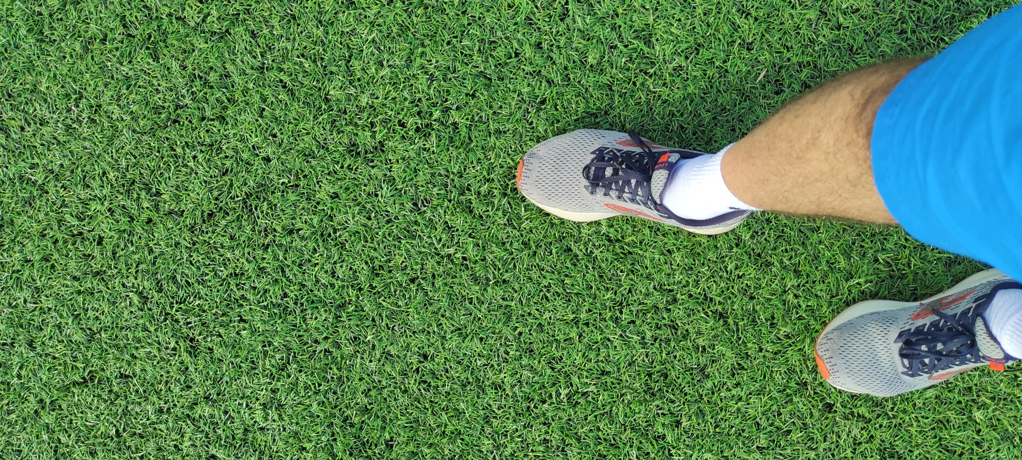 The image shows a top-down view of a person's legs and feet standing on artificial grass. The person is wearing grey running shoes with dark blue laces and orange accents, white socks, and blue athletic shorts. The artificial grass is green and appears to be densely packed. The scene does not provide any visual cues to determine the specific location in the city of Noviy, Russia. The image captures a static moment of someone standing, with no apparent action or interaction taking place.