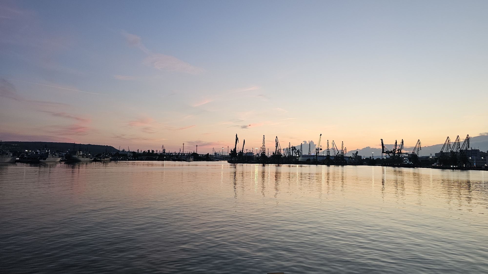 The image depicts a harbor scene in Varna, Bulgaria, at dusk. The sky displays a gradient of soft pinks, oranges, and blues, with wispy clouds. In the foreground, the calm water reflects the colors of the sky and the silhouettes of the structures on the opposite shore. The shoreline is lined with numerous industrial cranes, their tall, dark forms stark against the colorful sky. Several ships and boats are docked, visible as dark shapes. The overall atmosphere is serene and industrial, characteristic of a port city during sunset.