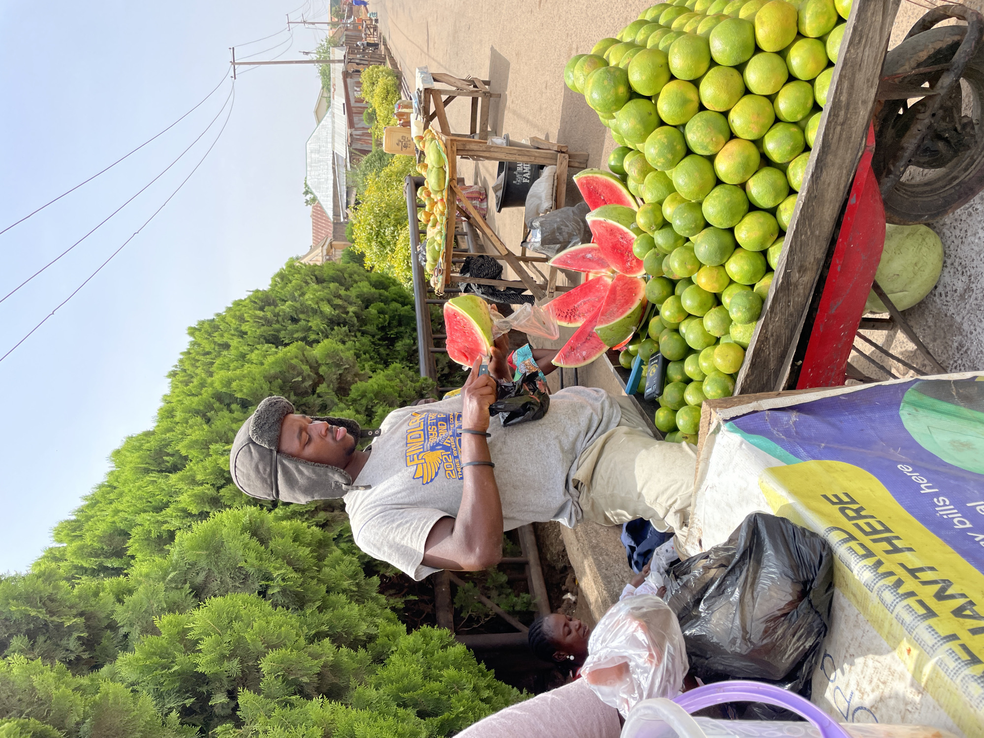 This image depicts a bustling outdoor fruit market scene in Jos, Nigeria, under a bright, clear sky. The central figure is a young man, dressed in a grey t-shirt featuring "FINDLAY", "2021", and "THREE RIVERS" text, light-colored pants, and a distinctive grey winter-style hat with earflaps. He is focused on handling a slice of watermelon, possibly cutting or preparing it for sale, with other vibrant red watermelon slices neatly arranged nearby.
Adjacent to the man is a large wooden cart overflowing with a considerable quantity of green citrus fruits, likely oranges or grapefruits, stacked high and ready for purchase. Behind this main display, a smaller wooden stall holds more fruits, including what appear to be papayas. A yellow jerrycan is also visible on this stall.
The market is set along a paved street or pathway, lined by dense, tall green conifer trees that create a rich natural backdrop. In the distance, simple buildings with corrugated metal roofs can be seen, along with utility poles and power lines stretching across the clear sky, indicating an active community setting. Various plastic bags, including a black trash bag and transparent shopping bags, are scattered around the base of the stalls. A partially visible banner or tarp in the foreground displays fragments of text such as "ANT HERE", "y bills here", "PERFECT", "HA", and "FAMI". Another individual is partially visible below the main subject, possibly sitting. The bright daylight suggests it is daytime, likely morning or late afternoon.