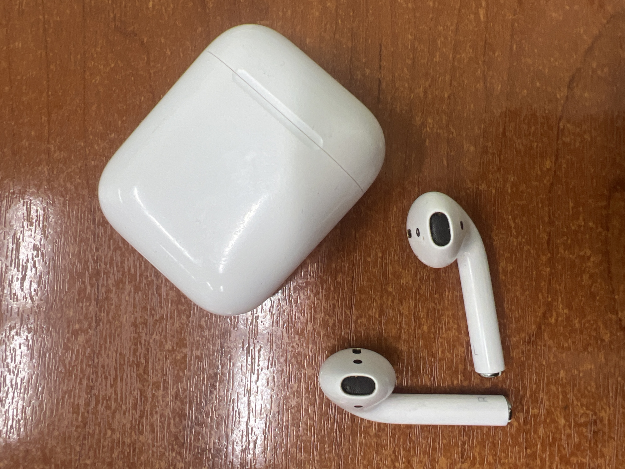 An overhead, eye-level shot shows a white charging case and two white wireless earbuds on a wooden surface. The charging case is positioned in the upper right quadrant of the frame, while the earbuds are in the lower left. One earbud lies horizontally to the right of the other, which is positioned vertically and slightly to the left. The wooden surface has a reddish-brown hue and a visible grain pattern, suggesting it is a table or desk. The scene appears to be indoors, and no specific location in Chaikovsky, Russia can be identified from the image.
