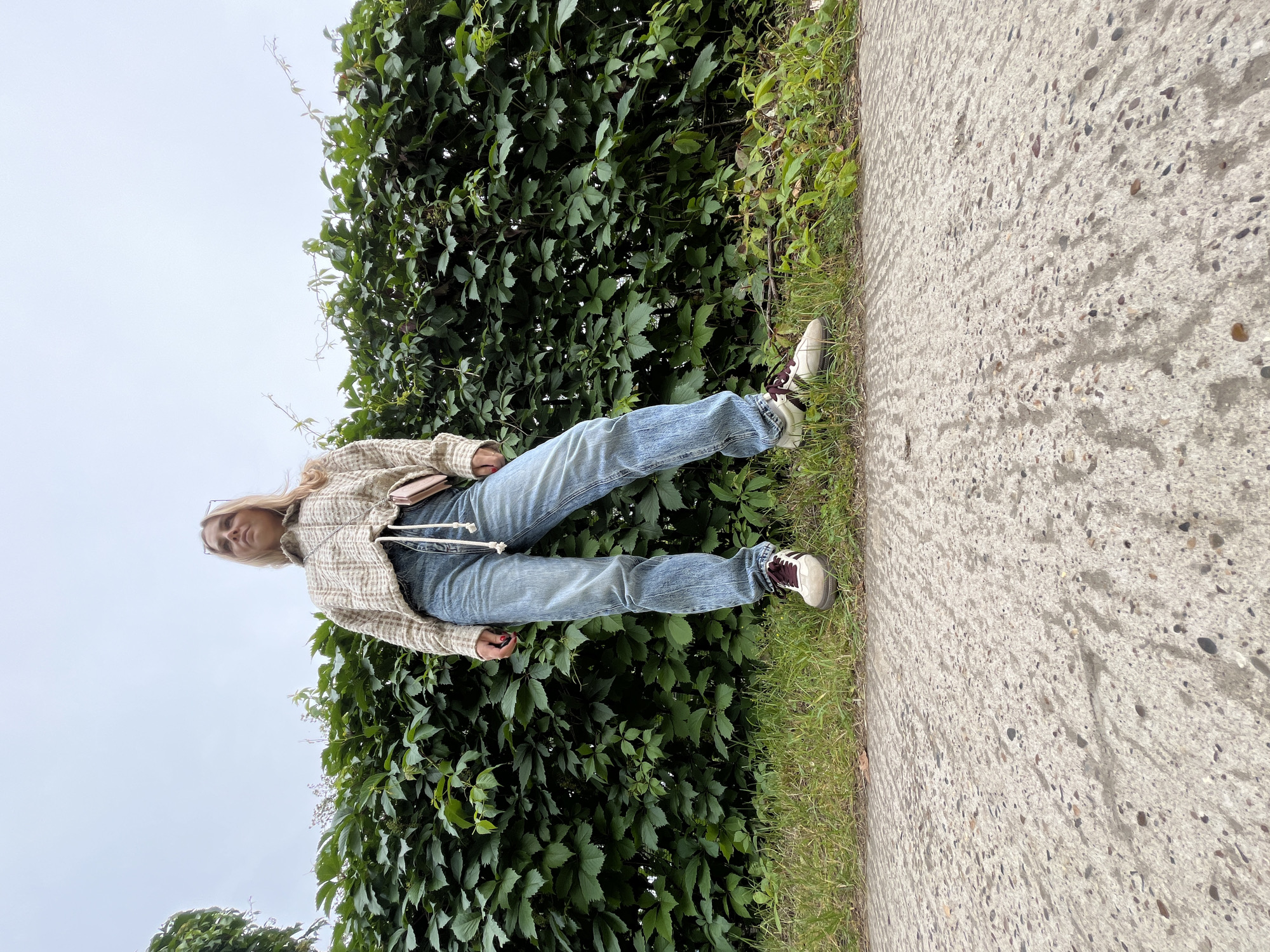 A low-angle medium shot captures a young woman standing on a concrete pathway in front of a dense, green ivy hedge. She is positioned centrally in the frame, with her body angled slightly to her left. She wears light-wash denim jeans, a plaid patterned collared jacket with a drawstring, and off-white sneakers with maroon accents. A small, pink cross-body bag is visible at her hip. Her blonde hair is shoulder-length and pulled back. The background consists of the lush green hedge and a pale, overcast sky. The foreground is dominated by the textured concrete path, with a strip of grass separating it from the hedge. The scene appears to be an outdoor setting, possibly in a park or along a street in the city of Chaikovsky, Russia.