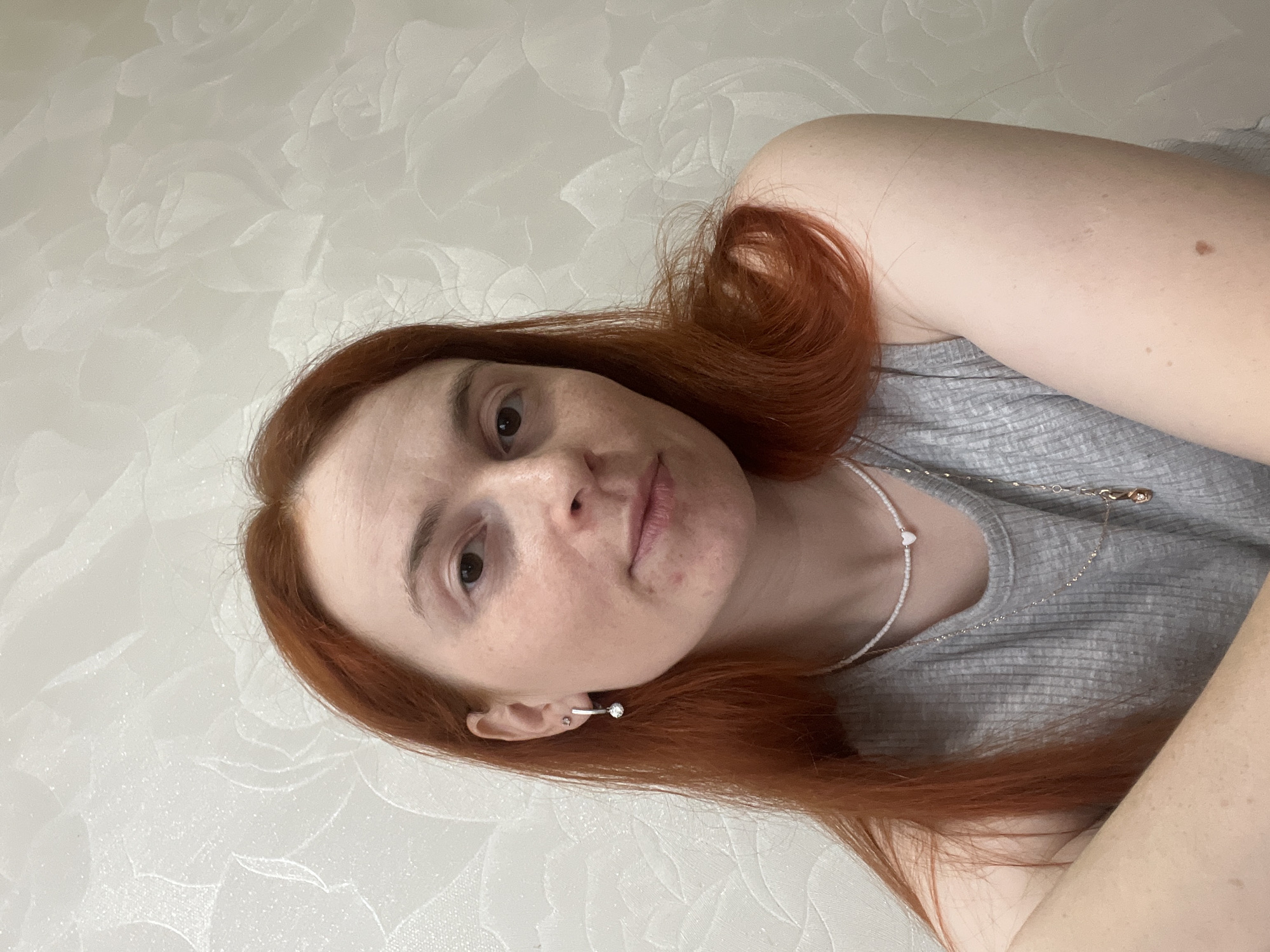 The image is a selfie of a woman with red hair and fair skin. She is wearing a gray shirt and two necklaces, one with small white beads and a heart charm, and another silver chain. She is wearing an earring in her left ear, which appears to be a curved silver bar with a gemstone at the end. The background is a textured off-white wall. The image was not taken in the city of Chaikovsky, Russia.