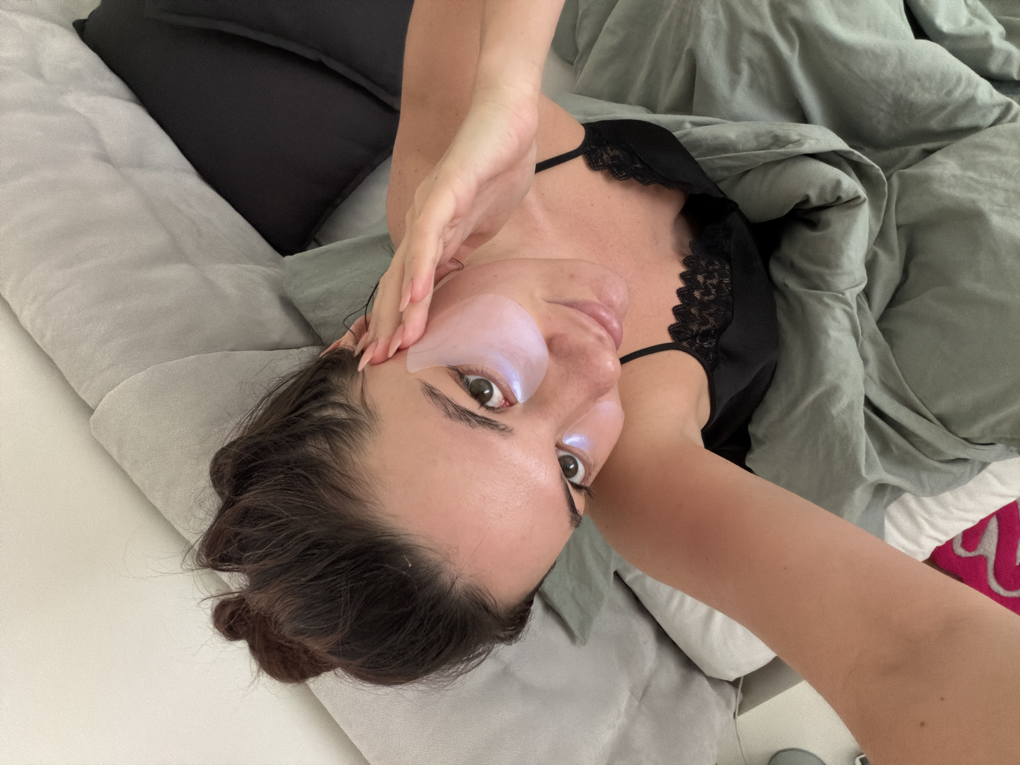 A woman is lying in a bed. She has dark hair, an eye mask on one eye, and is wearing a black lace top. She is partially covered by a gray duvet. There are various pillows in the bed, including one that is black and one that is gray. The bed has white sheets. The photo is taken from a top-down perspective. There is no indication of the location being Belgrade, Serbia.