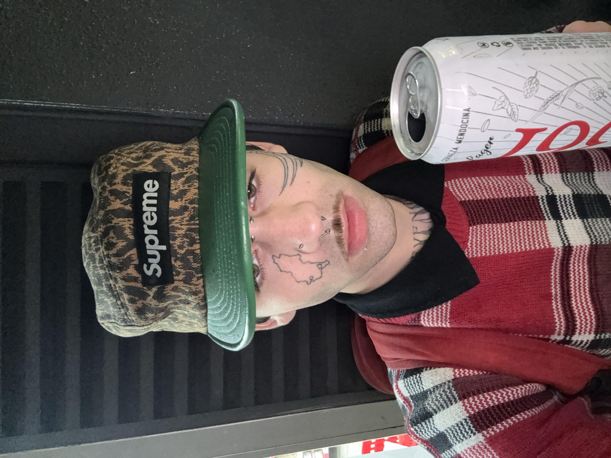 A close-up, sideways portrait features an individual, likely male, looking directly at the camera. The person wears a leopard print Supreme baseball cap with a green brim, and a red, black, and white plaid sweater or jacket. Their face is adorned with several piercings, including a hoop in the left nostril, two studs below the left eye, and a labret piercing on the lower lip. Distinctive facial tattoos include a map-like outline on the left cheek and a series of thin, curved lines above the right eyebrow. A tattoo is also partially visible on the neck.
An opened white beer can rests beside the person. The can's label displays illustrations of hops, wheat, and barley in silver, alongside the text "CERVEZA MENDOCINA" and "Lager." Large red lettering partially visible on the can appears to start with "JOL..." or "JOH...". The "Mendocina" reference links to Mendoza, an Argentine province, consistent with the Buenos Aires, Argentina location context. The background is a dark, textured surface with faint vertical stripes, contributing to the casual and informal setting. The image's sideways orientation suggests an unusual photographic angle.