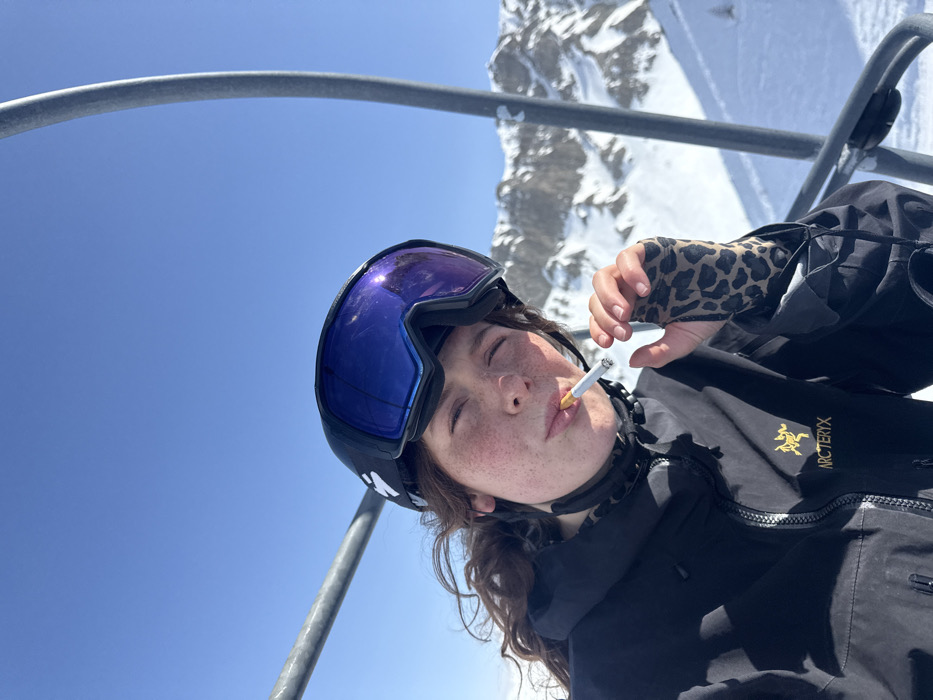 A female individual is photographed in a close-up, chest-up view, seated on a ski lift in Le Monêtier-les-Bains, France. She faces the camera with a slight upward gaze. Her head is covered by a black ski helmet with an unidentified white logo on the left side, and reflective blue-purple ski goggles are pushed up on the helmet. Dark, wavy hair is visible below the helmet. Her face shows freckles across the nose and cheeks. She wears a black ski jacket, identified by a yellow Arc'teryx logo on the left chest, and a leopard-print fingerless glove on her right hand. A lit cigarette is held between her pursed lips. The background consists of a clear blue sky and snow-covered mountain ranges under direct sunlight. Two gray metal ski lift poles are visible, framing the subject on the left and right sides of the image.