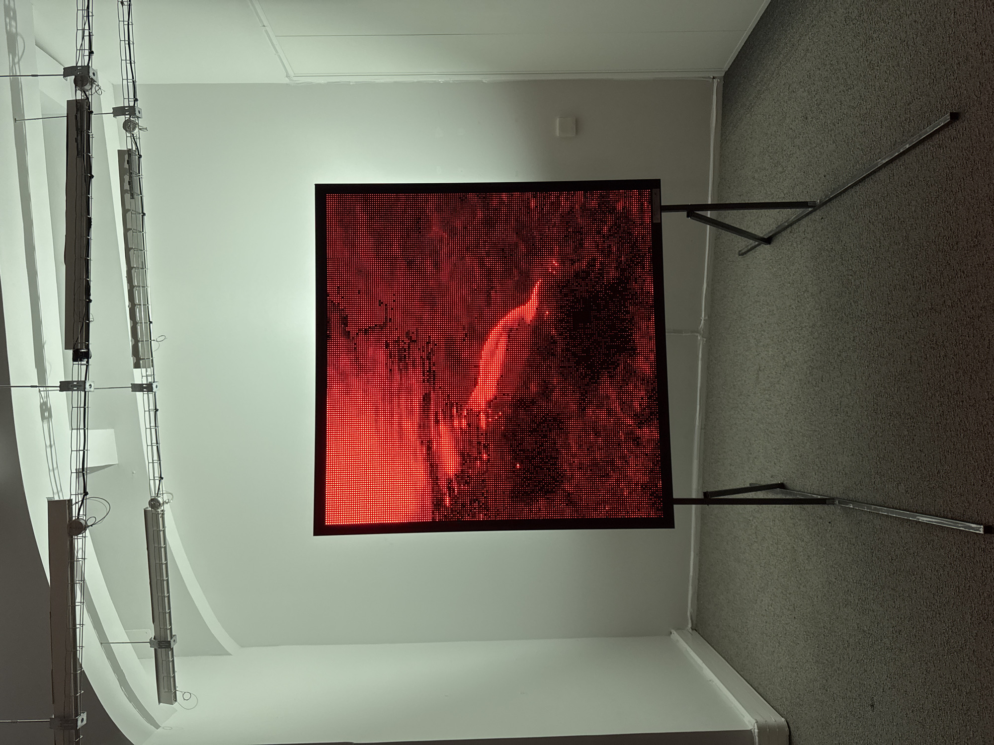 An indoor scene showcases a prominent, large square LED screen standing on a dark metal frame in what appears to be an exhibition or gallery space. The screen displays a highly pixelated, intense red image, which vaguely depicts a human-like form or torso amidst swirling, fiery textures. The digital art piece is the focal point, casting a warm red glow onto the surrounding area.
The room features light-colored, possibly white, walls and ceiling. On the left side of the image, two parallel vertical structures, resembling exposed metal frames or ladders, are mounted against the wall. These structures, possibly part of a lighting rig or cable management system, contain wires and cylindrical components. The floor is covered with a dark grey, textured carpet, and the screen's stand includes two angled supports extending onto this carpet. The overall ambiance is modern and clean, focusing attention on the illuminated display. No specific text is visible within the image.