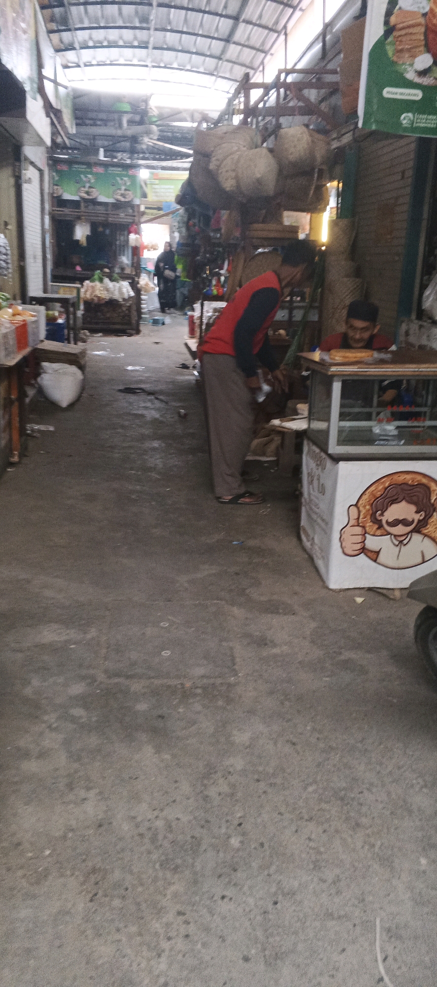 The image depicts an outdoor market scene in Sekaran, Indonesia. In the foreground, a person wearing a red and black long-sleeved shirt and grey pants is bending over near a stall. To their right, another person, presumably a vendor, is visible behind a glass-fronted display counter, wearing a black cap and a red shirt. The counter has a white box with a cartoon drawing of a person with a mustache giving a thumbs-up. The market aisle is paved with concrete and shows some debris. Overhead, a corrugated metal roof structure with supporting metal beams covers the market. Various goods, including sacks and what appear to be food items, are displayed at stalls along the sides of the aisle. Other individuals are seen further down the aisle, including a person in a black headscarf.