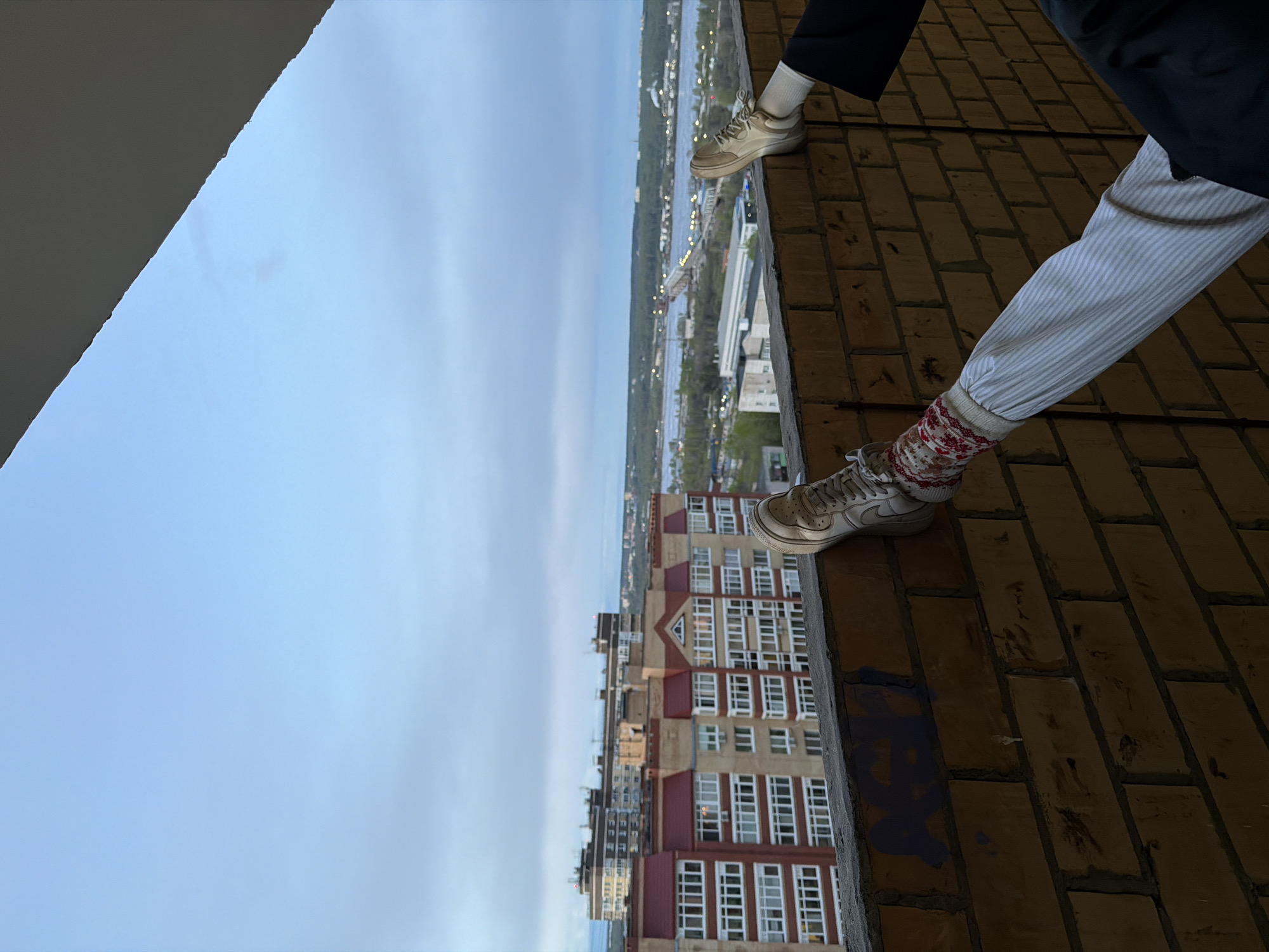 The photograph depicts two people standing on the edge of a brick building, with a view of the city of Perm, Russia, in the background. One person is wearing dark pants and white socks and light-colored sneakers, while the other is wearing light-colored pinstriped pants and festive red and white socks and light-colored sneakers. The scene includes the sky, other buildings, and a river.