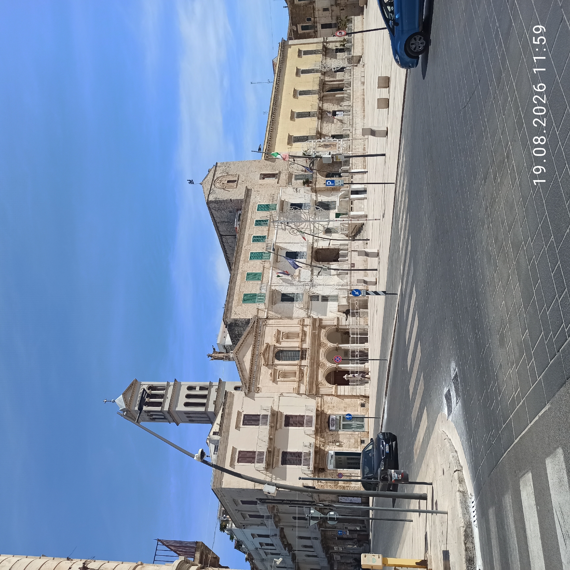 The image shows a view of Piazza Matteotti in Ruvo di Puglia, Italy, on a bright, sunny day. A black car is parked in the foreground, with a pedestrian crossing in front of it. Beyond the car, the piazza opens up to reveal several historic buildings, including the Chiesa Madre and the Palazzo Senatorio. The Chiesa Madre, on the left, features a tall bell tower and a facade with arched windows. The Palazzo Senatorio, to the right of the church, is a palatial building with a long facade and a yellow-toned exterior. Decorative white structures, possibly for a festival or event, are visible in the piazza. A blue car is parked on the right side of the image. The sky is clear blue with a few wispy clouds. The date and time stamp "19.08.2026 11:59" is visible in the bottom right corner of the image.