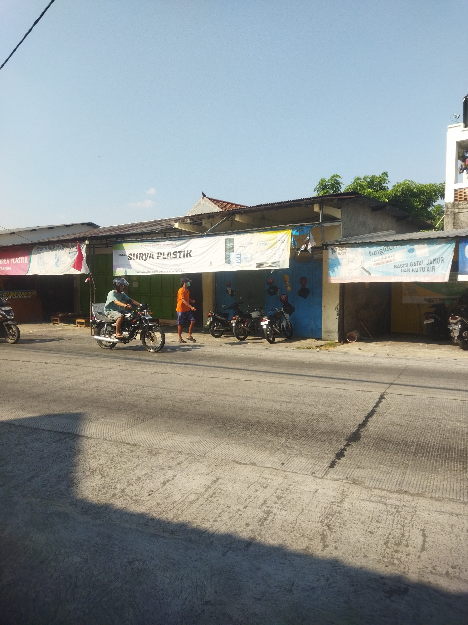The image depicts a street scene in Juwiring, Indonesia, likely in the afternoon given the shadows. The foreground shows a concrete road with a distinct shadow cast from the left side of the frame. In the middle ground, a row of storefronts lines the street. The buildings have corrugated metal roofs and are painted in various colors, including white, blue, and yellow. Several businesses are advertised with banners, including "SURYA PLASTIK" and a banner advertising "fungiderm" for skin conditions.
Several motorcycles are parked in front of the shops, and two men are visible. One man, wearing a light blue t-shirt, dark shorts, and a helmet, is riding a motorcycle. Another man, in an orange t-shirt and checkered shorts, is standing near the shops. A red and white Indonesian flag is displayed on a pole attached to one of the shops. Trees and overhead power lines are also visible in the background. The overall impression is that of a typical commercial street in a town.
