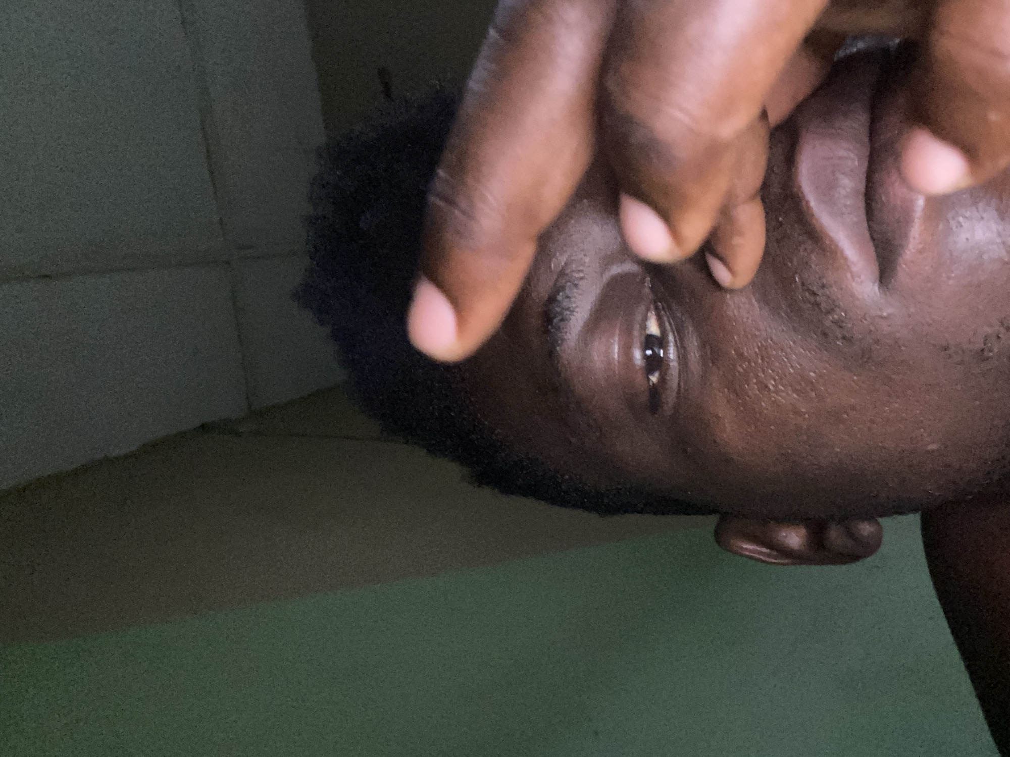 A close-up, rotated view of a dark-skinned person with short, dark hair. The person's hand is obscuring part of their face, with fingers near the eye and nose. The background consists of a wall, partially covered by what looks like flooring material. The scene location is Unknown, Unknown.