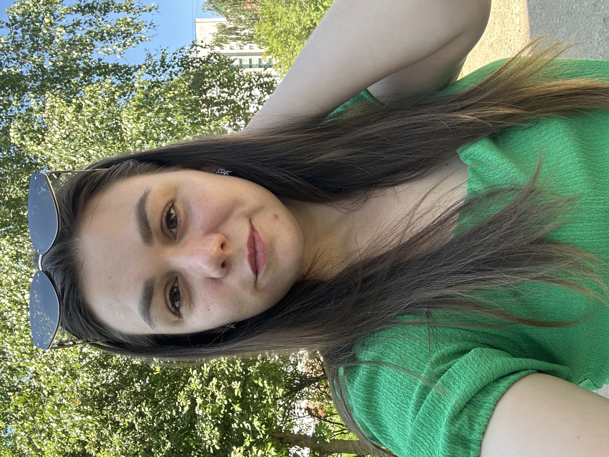 A young woman is taking a selfie outdoors with a background of trees and buildings under a clear blue sky. She is wearing a bright green top and has sunglasses perched on her head. Her left arm is raised, with her hand resting on the back of her head. Her hair is long and brown, and she is wearing small earrings. She has a light complexion and is looking directly at the camera with a slight smile. The environment suggests a sunny day, possibly in a park or residential area in Chaikovsky, Russia, given the presence of trees and apartment buildings. The lighting indicates it's daytime.