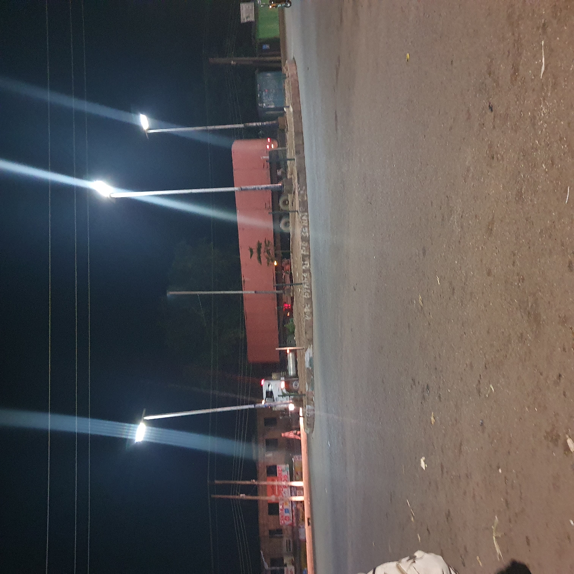 This image captures a nighttime scene in Bukuru, Nigeria, likely at a roundabout or intersection. A long, red truck trailer is prominently parked, its lights illuminated. In the background, several streetlights cast bright beams into the dark sky, creating streaks of light. A white truck is visible to the left of the trailer, and a few people appear to be near the vehicles and buildings in the background. The ground is paved, suggesting a road or public area, and there is some debris scattered on the surface. The overall atmosphere is industrial and utilitarian, with the artificial lighting highlighting the vehicles and infrastructure.