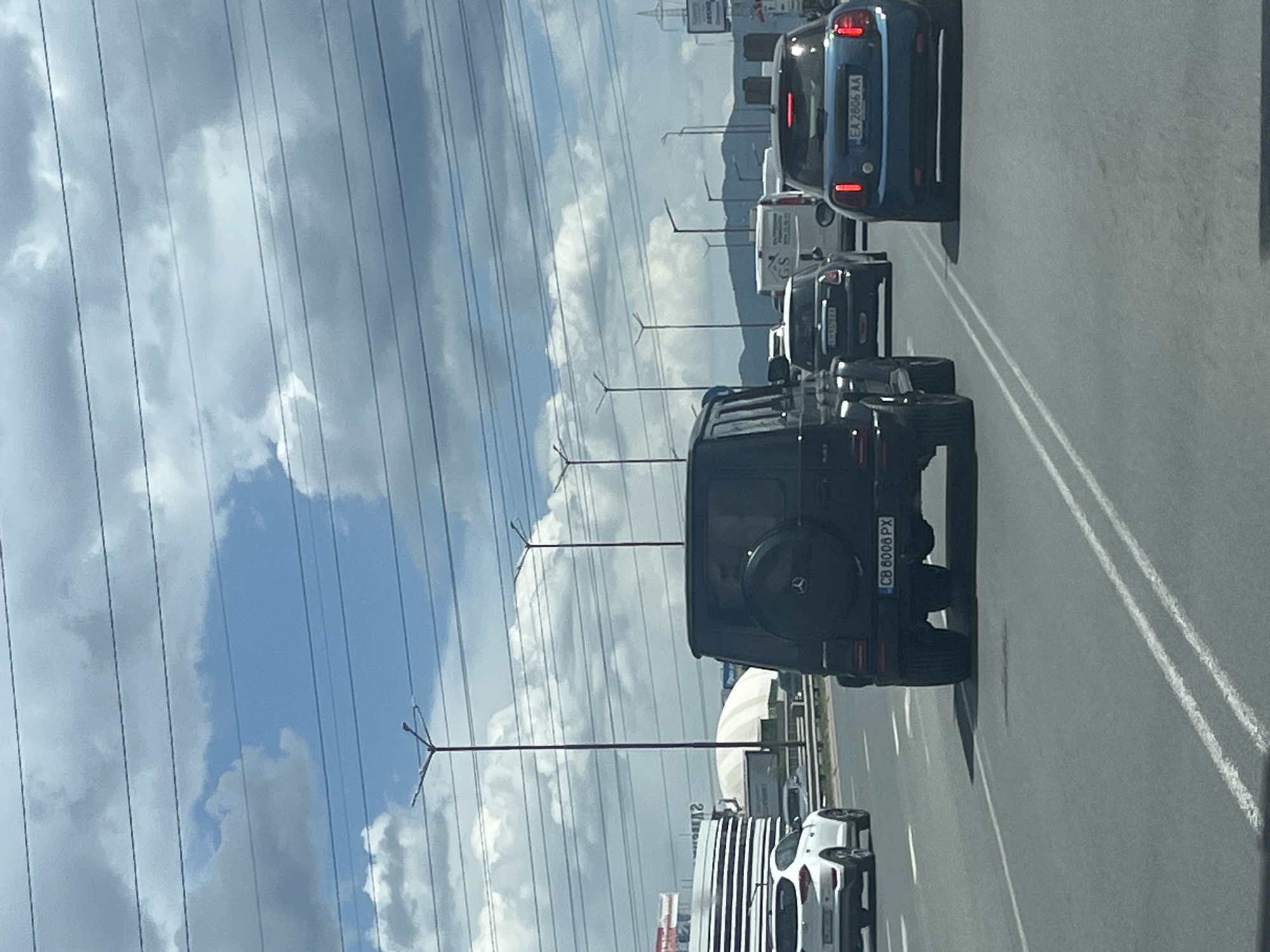 The image shows a traffic scene in Sofia, Bulgaria. Cars are in multiple lanes on a highway or major road. A black Mercedes-Benz G-Class with the license plate "CB 8008 PX" is prominently featured. Other vehicles include a white car, a grey SUV, and a blue car with the license plate "EA 2806 AA." Overhead power lines and streetlights are visible. In the background, there are buildings and clouds. The sky is partly cloudy.