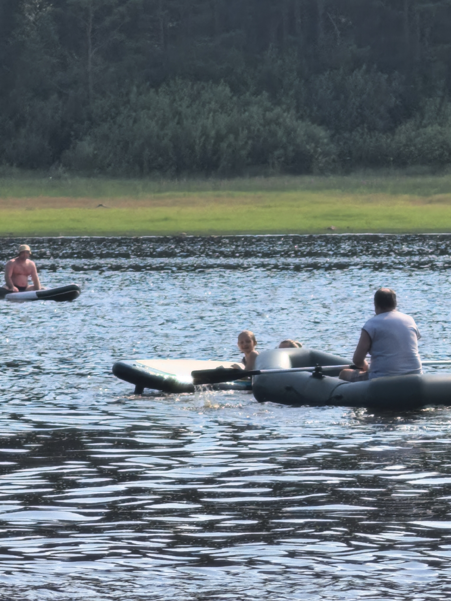 In Sysert, Russia, on a bright, sunny day, three individuals are enjoying recreational activities on a tranquil body of water. In the foreground, a man is seated in a green inflatable boat, facing away from the camera and appearing to be steering. Beside him, a young child with light-colored hair is visible, looking towards the camera. Another inflatable object, possibly a raft or a second boat, is partially submerged in the water in the center foreground, with its dark sides and a light-colored top visible. In the mid-ground, a man is sitting on a black and white inflatable raft, facing left. The background features a dense, dark green forest that stretches across the horizon, meeting a strip of bright green grass and a darker line of rocky shore along the water's edge. The water is a deep blue, with numerous ripples and reflections of sunlight shimmering on its surface.