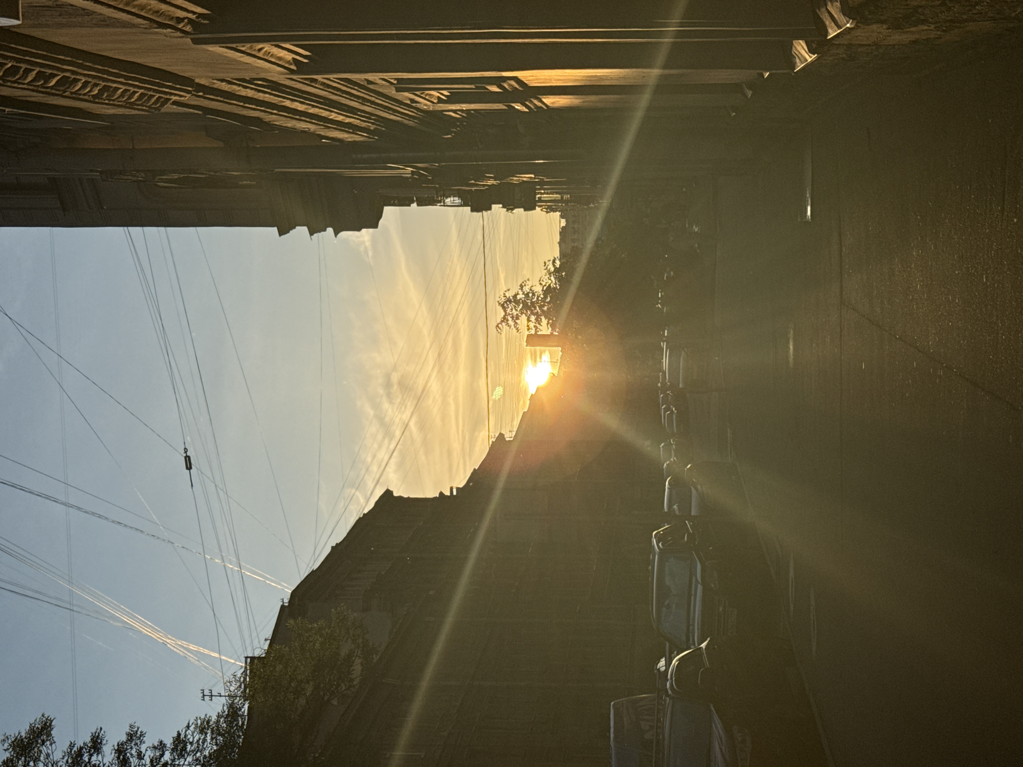 This is a medium shot, vertical, low-angle photograph of a street in Saint Petersburg, Russia during sunset. The sun is low in the sky, casting long shadows and creating lens flare across the image. The street is lined with cars parked on both sides, and vintage-looking buildings with ornate facades. In the background, there are telephone wires crisscrossing the sky. The sky is a pale blue with streaks of white clouds. The image is dark due to the low light conditions.