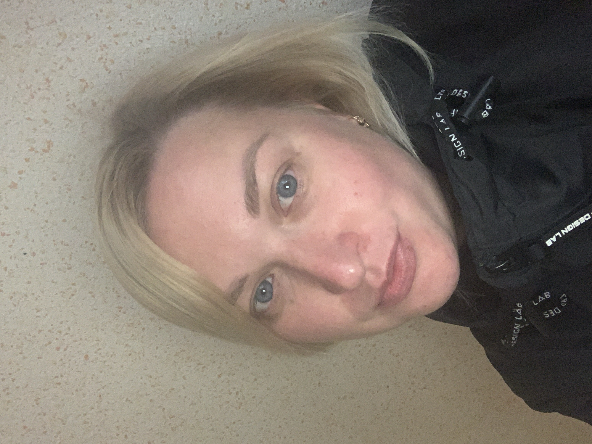 A close-up self-portrait features a woman with blonde hair and blue eyes, captured indoors in Chaikovsky, Russia. She looks directly at the camera with a neutral to slightly smiling expression. Her skin appears fair with some natural redness on her cheeks and nose. She is wearing a black jacket or hoodie, and a small gold-colored earring is visible in her right ear. The background consists of a light, speckled surface, possibly a wall or countertop. Visible text on the drawstring of her jacket repeatedly spells out "DESIGN LAB". The lighting is soft and even, suggesting an indoor setting.