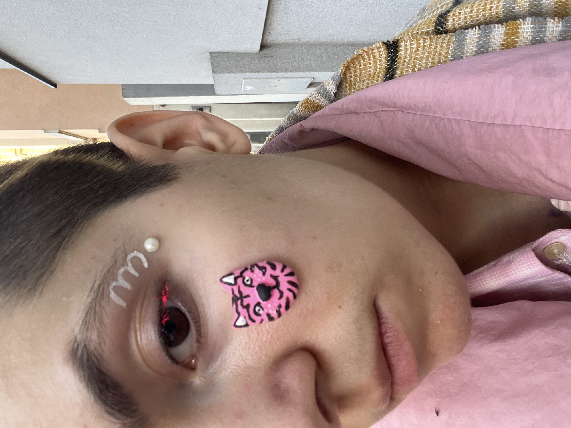A close-up, rotated portrait shows a person's face decorated with artistic embellishments. Above the left eye, a white, wave-like design is adorned with a pearl-like bead. A pink tiger-shaped sticker is positioned near the right eye. The person is wearing a pink top and a plaid jacket is partially visible. The setting is likely indoors given the ceiling in the background. There is no information in the image to specify the location as the city of Belgrade, Serbia.