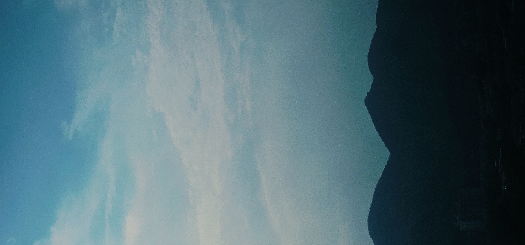 The image depicts a dark silhouette of mountains against a gradient sky of pale blue and white clouds. In the lower portion of the image, a faint outline of buildings and structures can be discerned at the base of the mountains, suggesting an urban or semi-urban environment. The overall scene is dark and moody, with the sky occupying the majority of the frame. The location is identified as the Chongqing High-tech Zone, China.