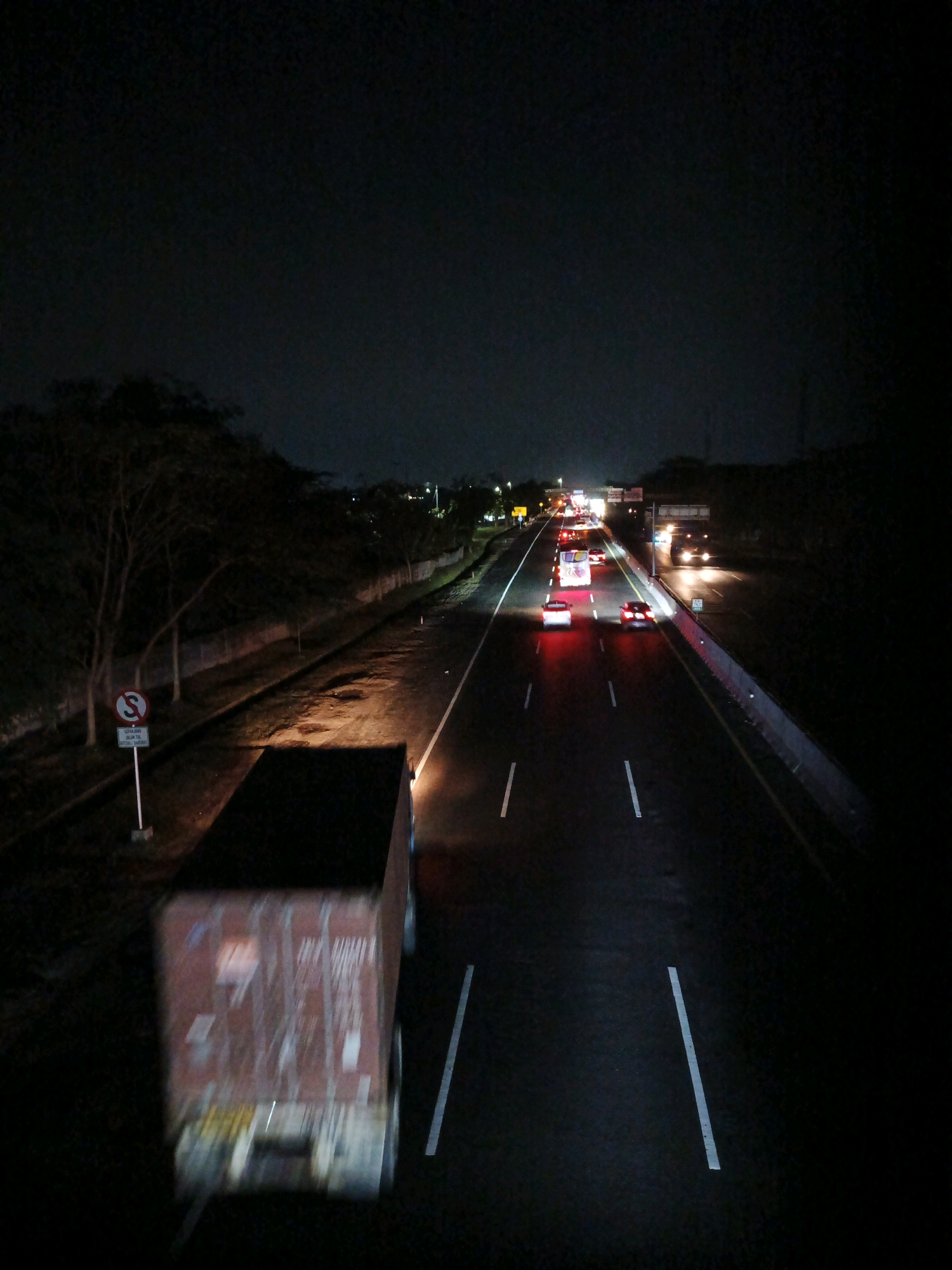 The image depicts a dark, nighttime scene on a highway, likely located in or near Kludan, Indonesia. The highway is multi-lane and appears wet, with reflections of vehicle lights on the surface. Several vehicles, including cars and a truck, are visible, their headlights and taillights illuminating the road. The truck in the foreground is partially blurred due to motion. On the left side of the highway, there is a tree-lined embankment with a concrete wall and a signpost. The sky is dark and overcast, with minimal visible features. The overall impression is of a busy highway at night, with limited visibility.