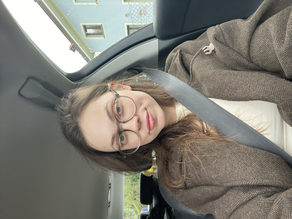 A young adult female is depicted seated in the front passenger seat of a vehicle, looking directly at the camera. She has medium-length brown hair and wears round, clear-framed glasses with dark temples. Her lips are slightly upturned, with a visible pink hue. She is dressed in a light-colored, possibly off-white, ribbed top, covered by a dark brown herringbone-patterned coat. A grey seatbelt crosses her torso diagonally from her left shoulder to her right hip. A small, light-colored, multi-pronged object is pinned to the right lapel area of her coat.
The vehicle interior shows a grey headliner and dark trim. Through the passenger window, a light blue building with a reddish-brown tiled roof and two dark-framed windows is visible. A light-colored metal fence with a partially obscured red and white logo (resembling a Shell symbol) is situated in front of the building. Raindrops are visible on the exterior of the car window, indicating precipitation. Green foliage is visible through the driver's side window. The scene is located in Monsummano Terme, Italy.