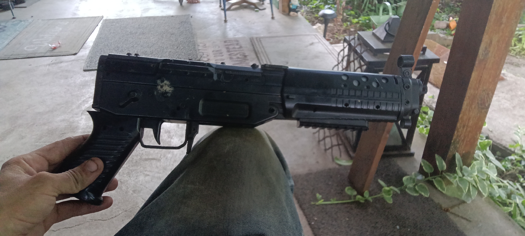 A person is holding a black airsoft gun. The person is sitting outdoors on a porch or patio with wooden posts visible. The location is Coal Hill, United States of America.