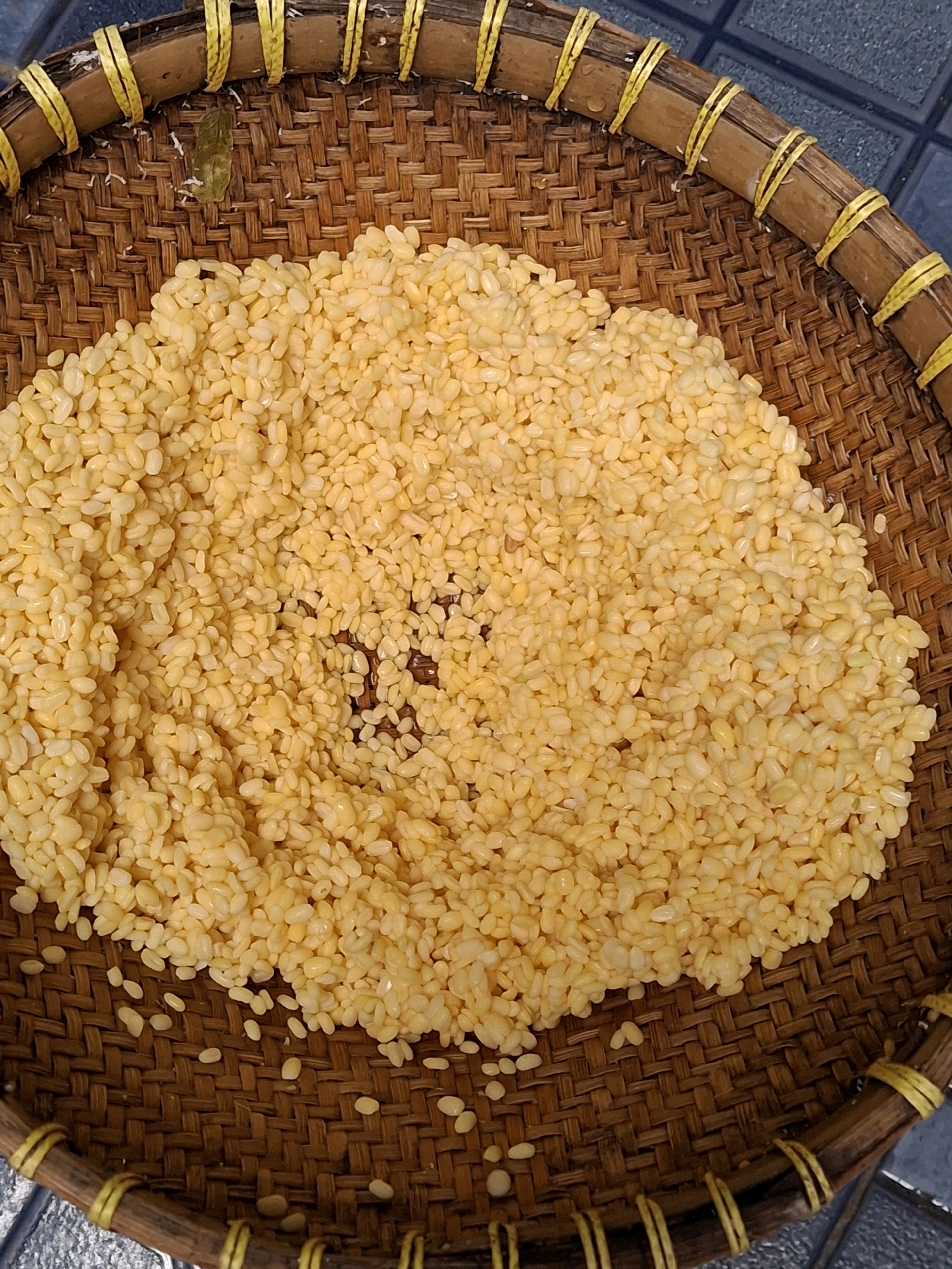 A close-up, top-down shot shows a woven bamboo tray filled with split yellow mung beans. The tray has a woven pattern with brown bamboo strips and is reinforced around the rim with yellow twine. The beans are primarily concentrated in the center of the tray, with a few scattered on the surrounding woven surface. The background is a dark, textured surface, possibly a tiled floor, which is out of focus. There are no people visible in the image. The scene is likely found in a home or market setting in Lumajang, Indonesia, where mung beans are a common food ingredient.