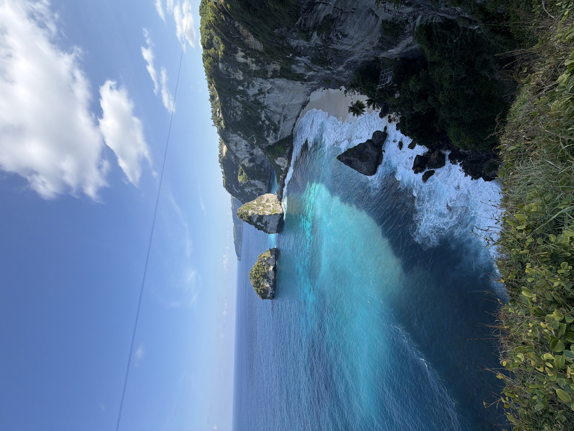 This is a landscape photograph taken from a high vantage point overlooking a tropical coastline in Nusa Penida, Indonesia. The scene features dramatic cliffs that plunge into the vibrant turquoise and deep blue ocean. Two large rock formations, covered in green vegetation, rise out of the sea in the middle distance. A small, sandy beach is visible nestled at the base of the cliffs. Gentle waves crash onto the shore, creating white foam. Lush green vegetation, including trees and grasses, covers the cliff tops and the foreground of the image. A thin wire or cable is visible stretched across the clear blue sky. The lighting suggests it is daytime, with sunlight casting shadows on the water and highlighting the textures of the cliffs. There are no people visible in the image.