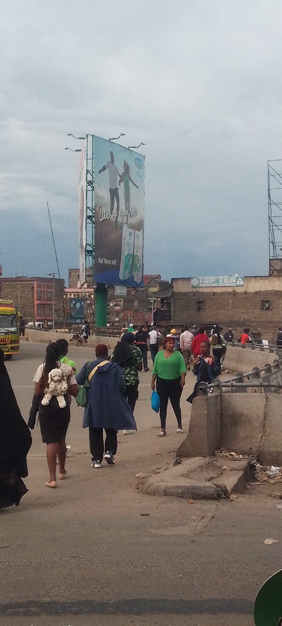 This image captures a busy street scene in an urban area, likely in Nairobi, Kenya, under an overcast sky which provides dull, diffused lighting. The scene is filled with pedestrians moving in various directions, suggesting a typical day's activity.
In the foreground and midground, several people are walking. One woman, seen from behind, wears a black dress and a distinctive white backpack shaped like a spotted dog. Another woman, facing partially towards the camera, wears a bright green top and black leggings, carrying a blue bag. A person in a blue jacket also walks with their back to the viewer. To the left, the front portion of a yellow and green public transport vehicle, characteristic of a Kenyan matatu, is visible.
Dominating the midground is a large, vertical billboard. The advertisement on the billboard features a man and a woman with outstretched arms, and text that reads "Lite up your life" and "Full flavor milk." Below the main image, graphics of milk cartons are displayed.
In the background, behind the street level, various urban buildings are visible, some appearing worn or under construction. On one of these buildings, a sign clearly reads "TOP TEN DRIVING SCHOOL." Concrete barriers line the edges of the pedestrian path, and some debris is scattered on the ground. The overall impression is one of a functional, active urban environment with ongoing daily life.