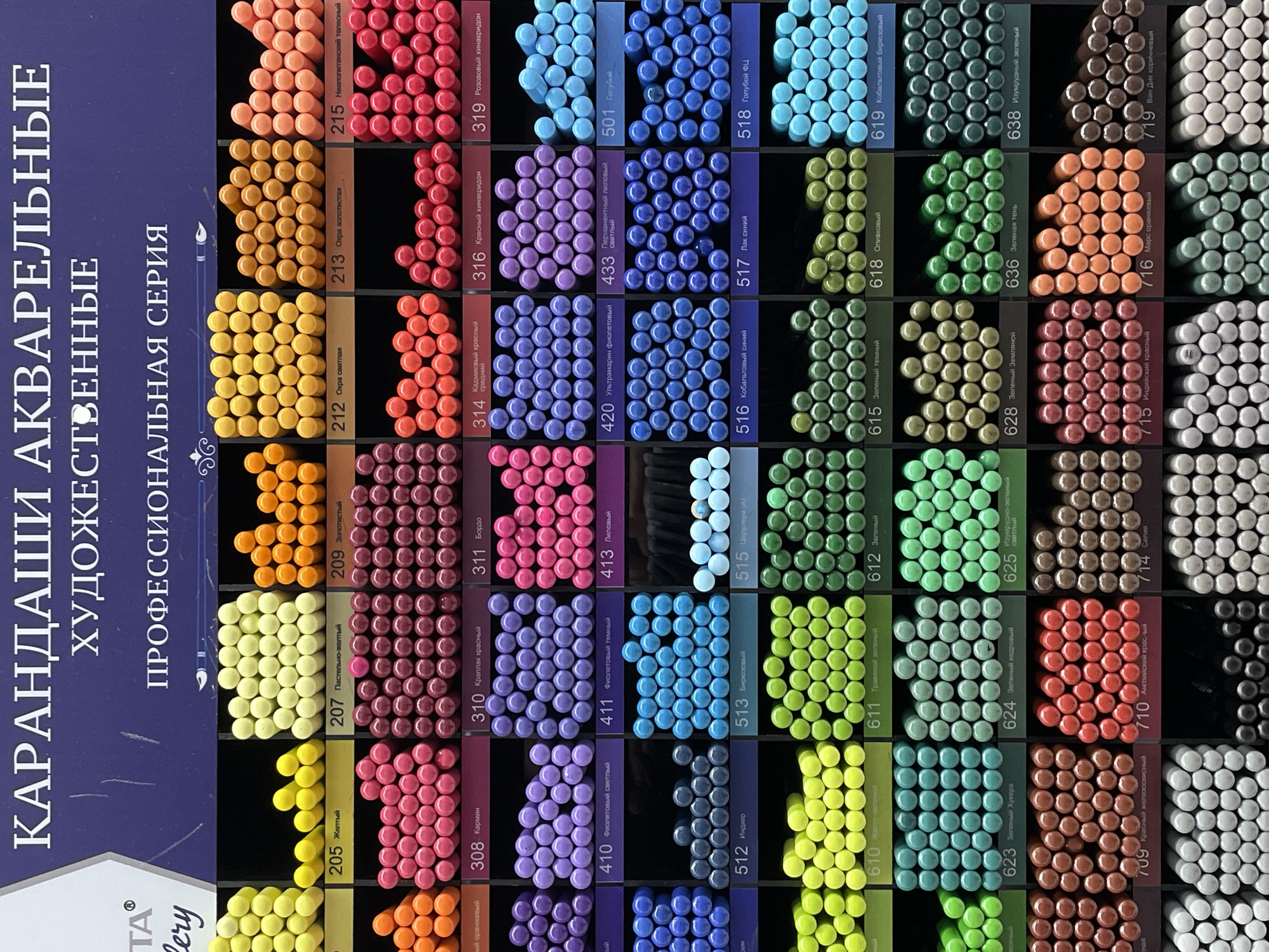 The image shows a display of watercolor pencils. Each pencil is arranged in rows and columns, grouped by color. Above each group of pencils is a label with a number and a description of the color in Russian. The image also shows a section of a blue box with text in Russian: "Карандаши акварельные художественные" (Watercolor pencils for art), "Профессиональная серия" (Professional series). The brand name "ТА gallery" is also visible. The color palette includes shades of yellow, orange, red, purple, blue, green, and brown.