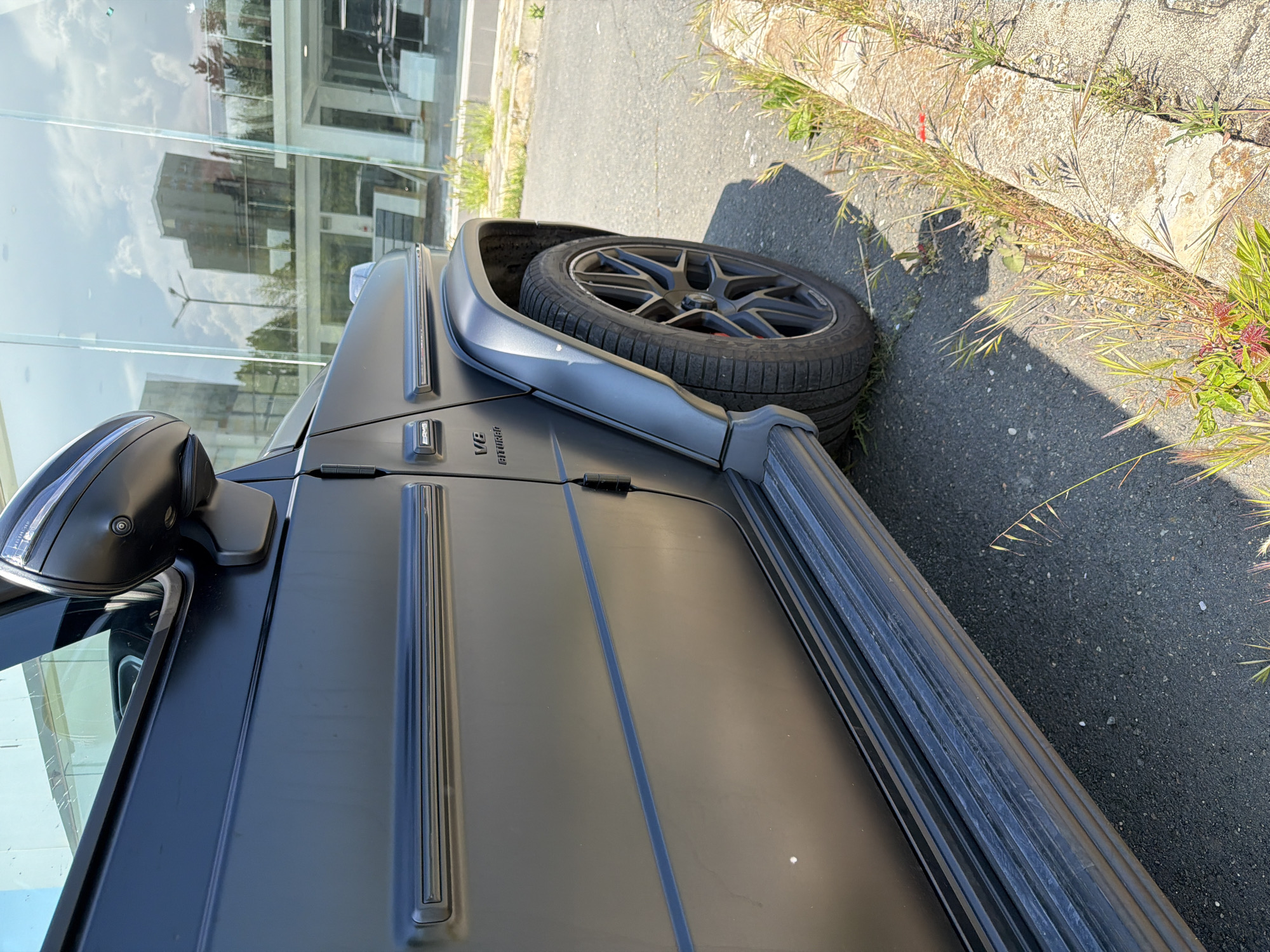 The image shows a portion of a matte black Mercedes-Benz G-Class parked on asphalt next to a raised concrete edge with vegetation. The vehicle's features visible include the front side mirror, part of the hood, the front door, the V8 BITURBO badging, a side step, and the front wheel. The wheel has black rims and a tire. A building with glass windows that reflect the sky and surrounding buildings is in the background. The location is Burgas, Bulgaria.